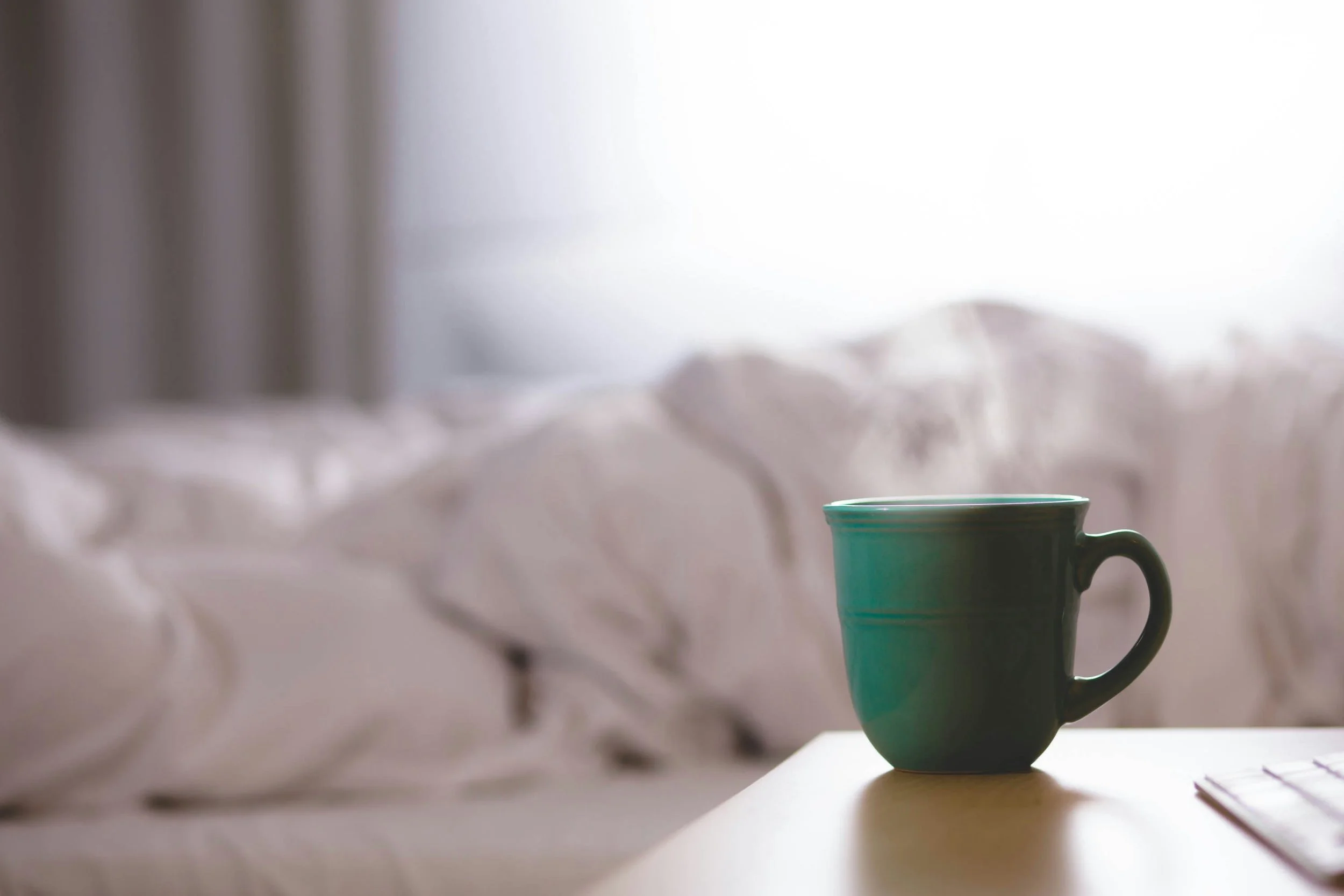 A teal mug on a wooden table near a white bed with rumpled sheets, in a softly lit room.