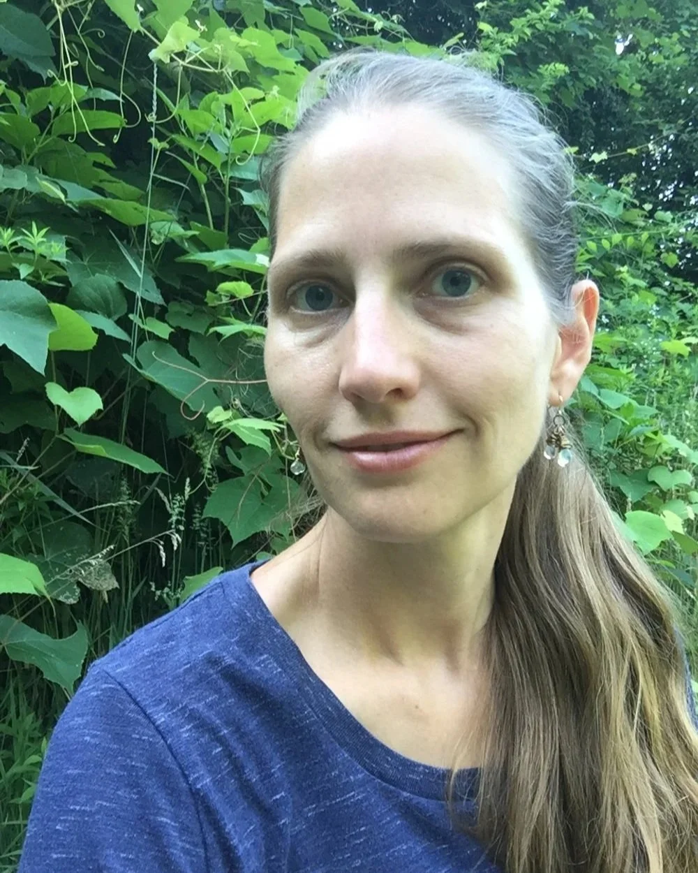 A woman with long brown hair and blue eyes smiling outdoors, surrounded by green plants and vines.