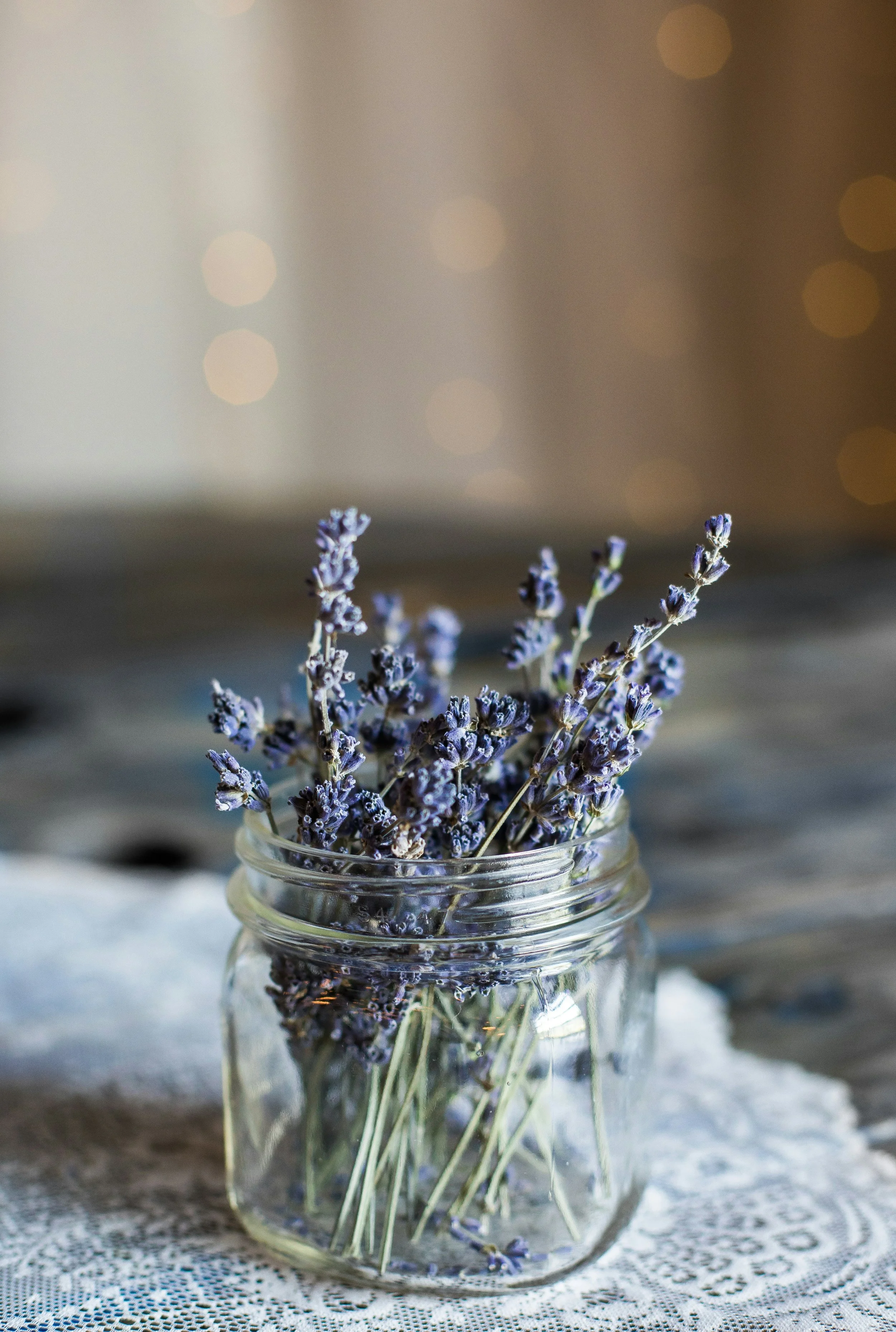 A cluster of purple lavender flowers in a clear glass jar placed on a lace cloth surface.