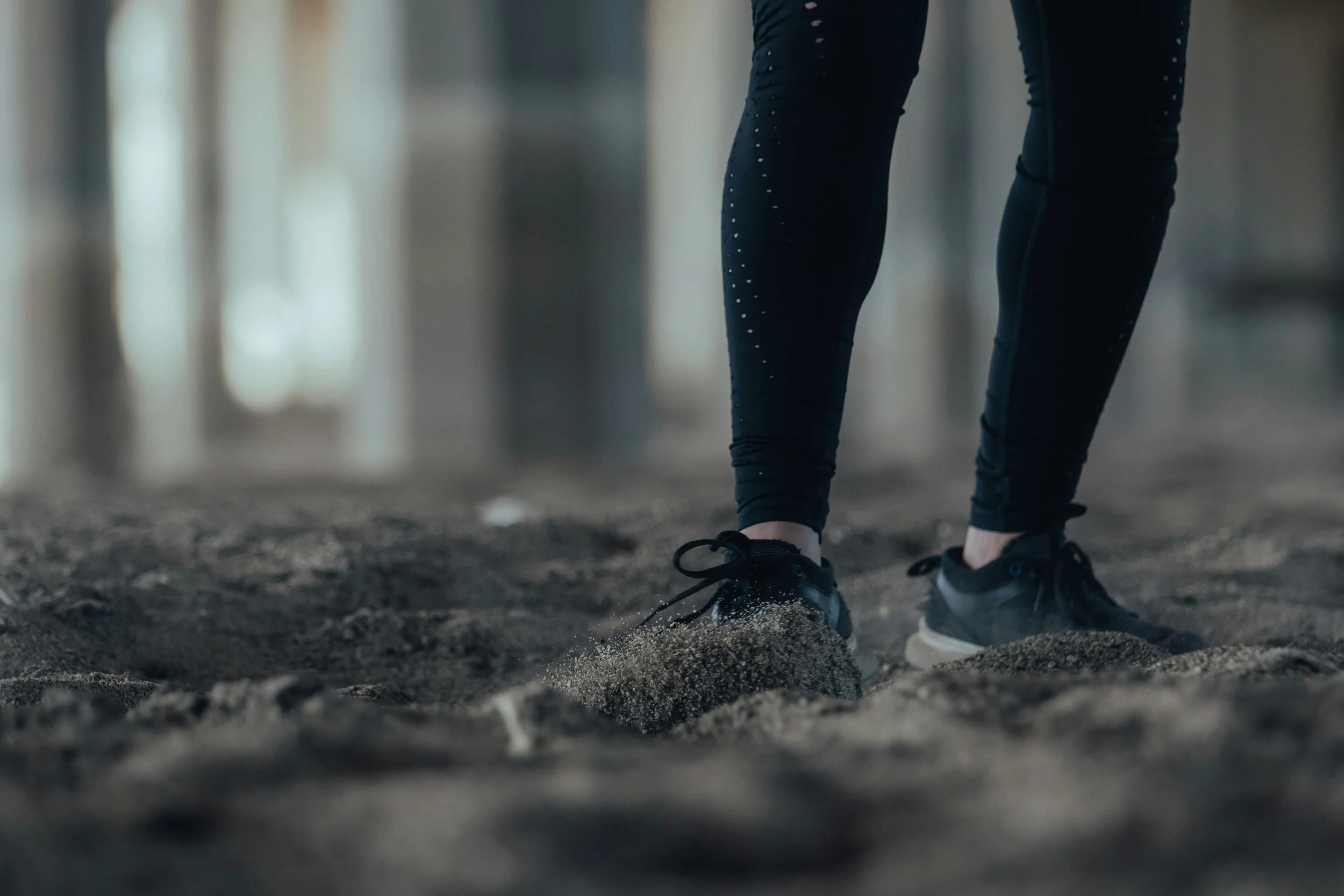Close-up of a person’s legs and feet in athletic shoes walking on sand, possibly on a beach or sand trail, with a blurred background.