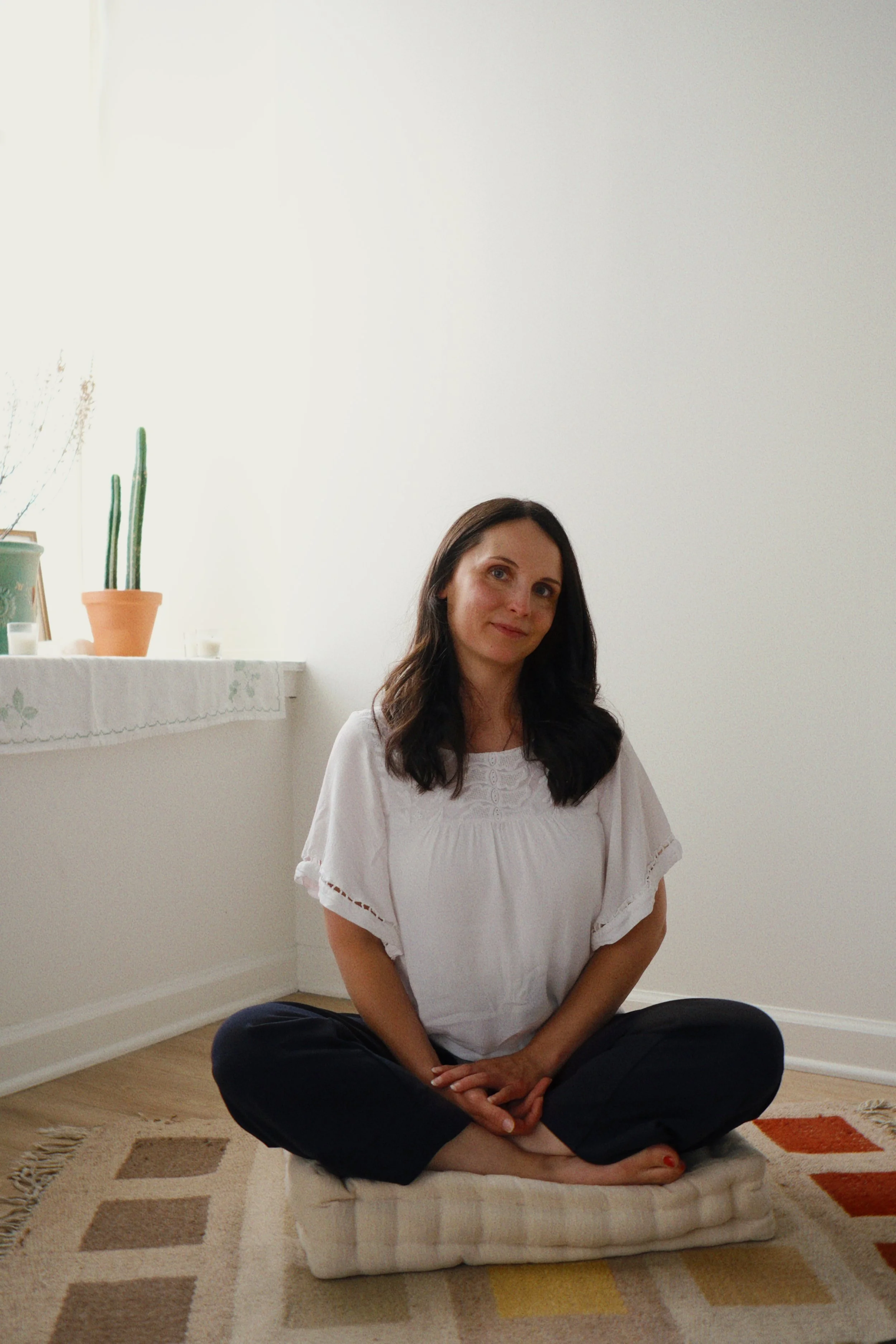 A woman with long dark hair, wearing a white top and black pants, sitting cross-legged on a cushion on a patterned area rug in a minimally decorated room with white walls. Behind her is a shelf with potted plants and candles.