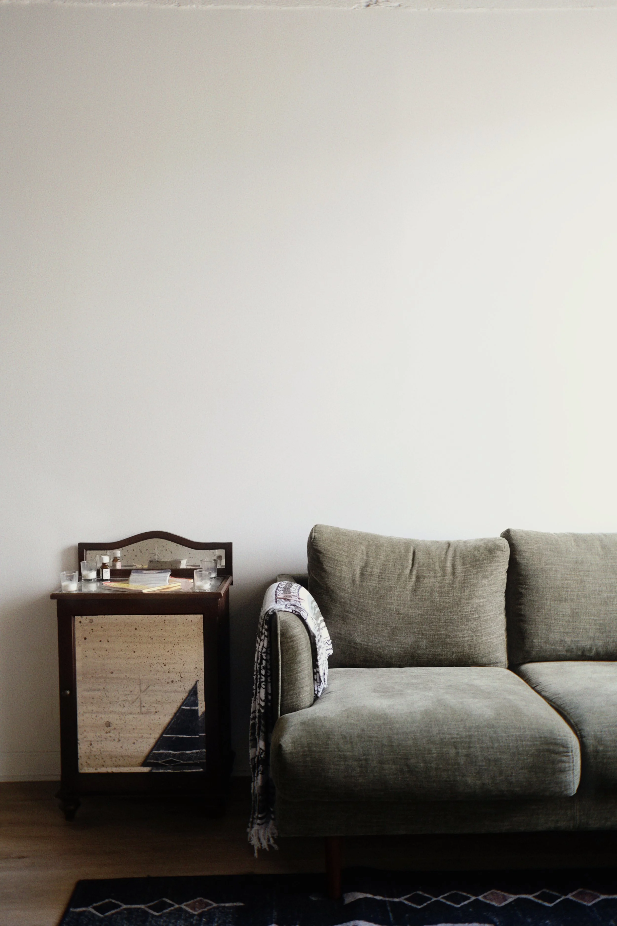 A living room corner featuring a gray upholstered sofa, a black and beige cabinet with glass jars and books on top, and a black rug with a white geometric pattern on a wooden floor. The wall behind is plain and light-colored.