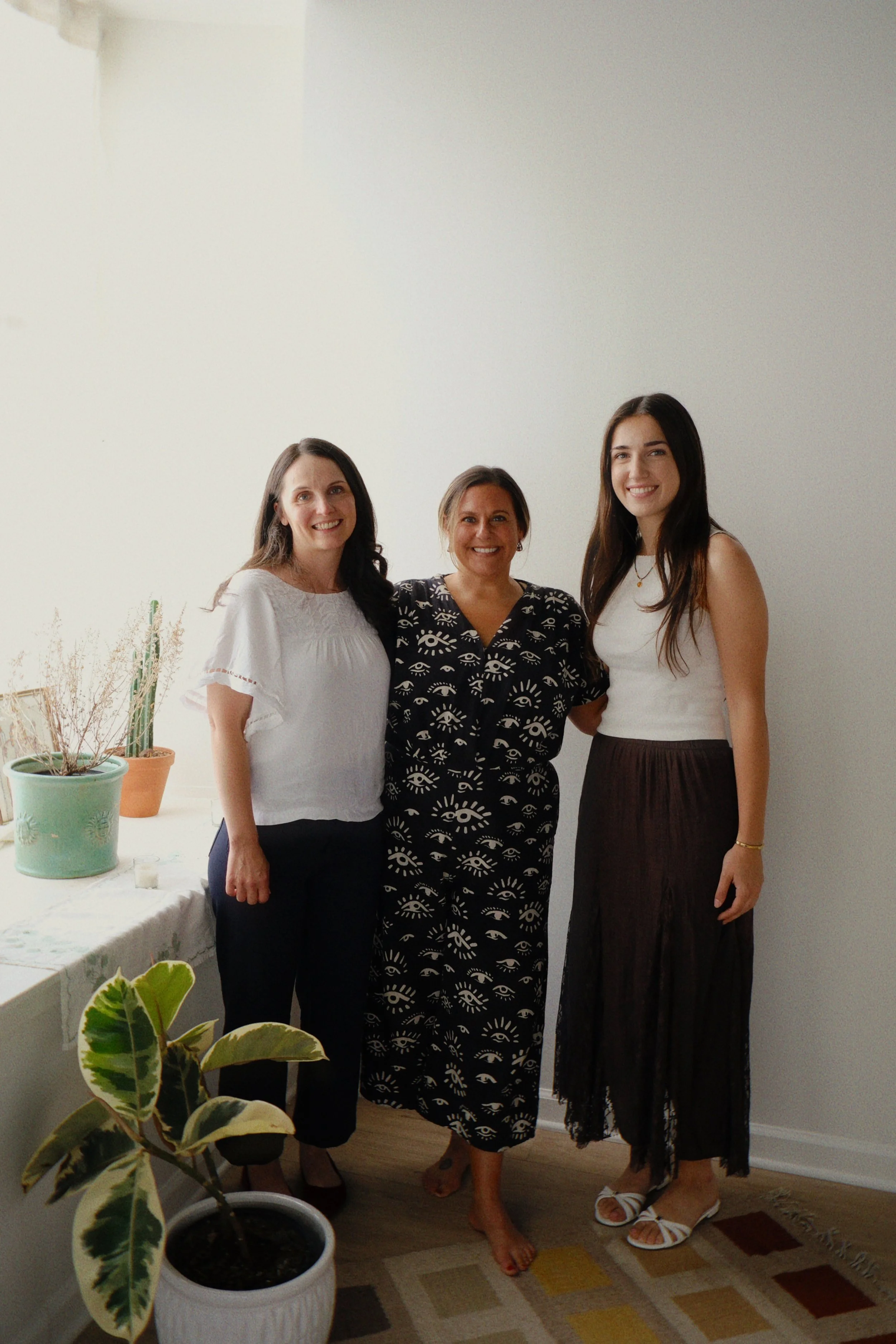 Three women standing together indoors, smiling for the photo. Behind them is a white wall, and to the side, a table with potted plants, including a variegated leaf plant and dried plants in a terracotta pot. The women are dressed casually, and a colorful geometric rug is on the floor.