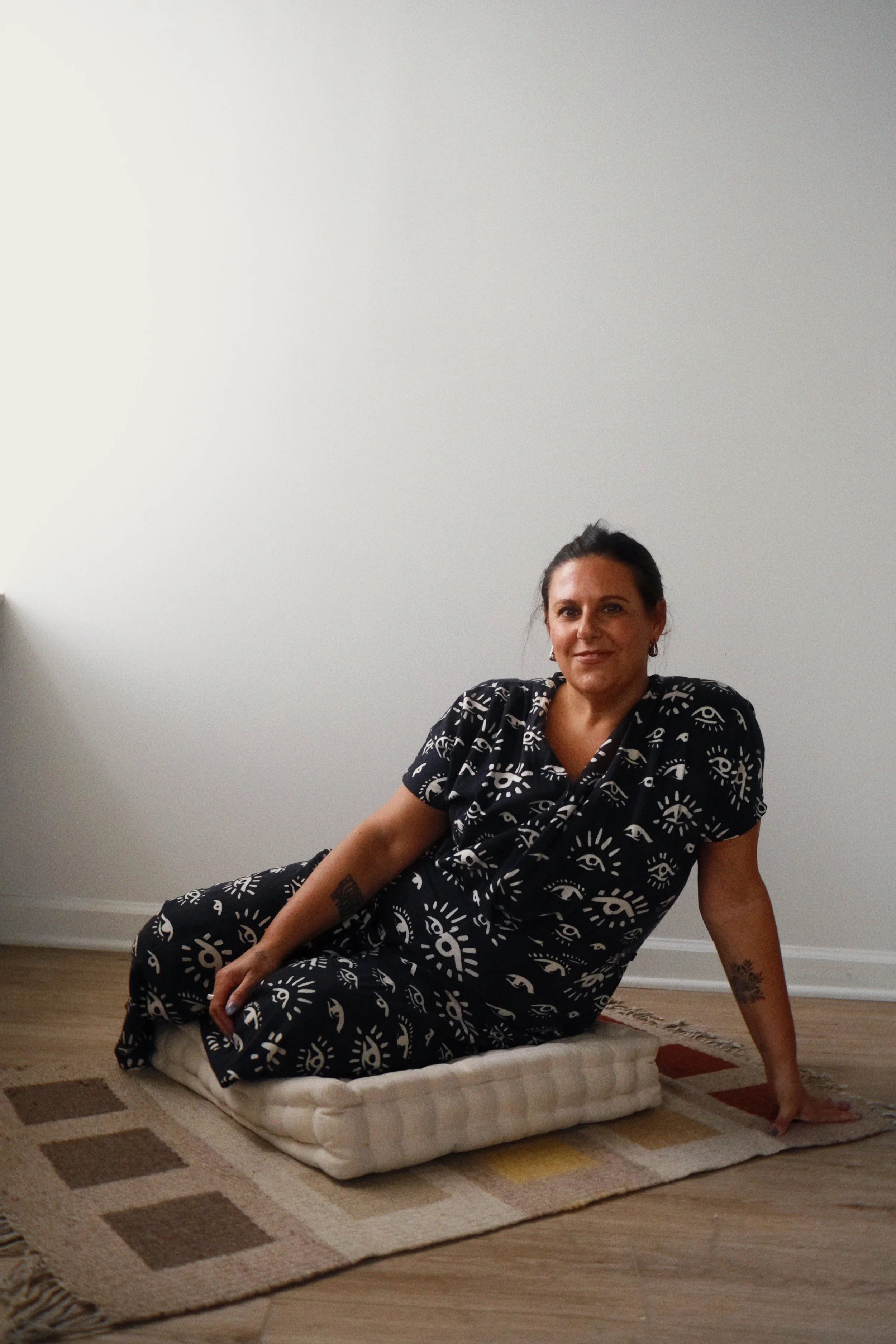 A woman in a black dress with white eye and sun pattern is sitting on a mattress on the floor, leaning back on one hand and smiling toward the camera, in a room with a plain white wall and a patterned rug.