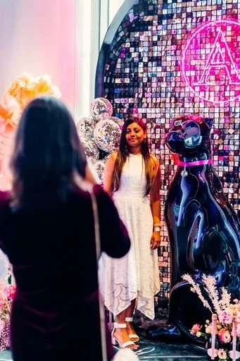 Two women are in a decorated space with shiny silver balloons, pink flowers, and a mirrored mosaic wall. One woman, with long dark hair, is facing away from the camera, while the other woman, with long brown hair, is facing the camera and smiling. There is a large black cat sculpture and a neon pink circular sign with some writing on the wall.