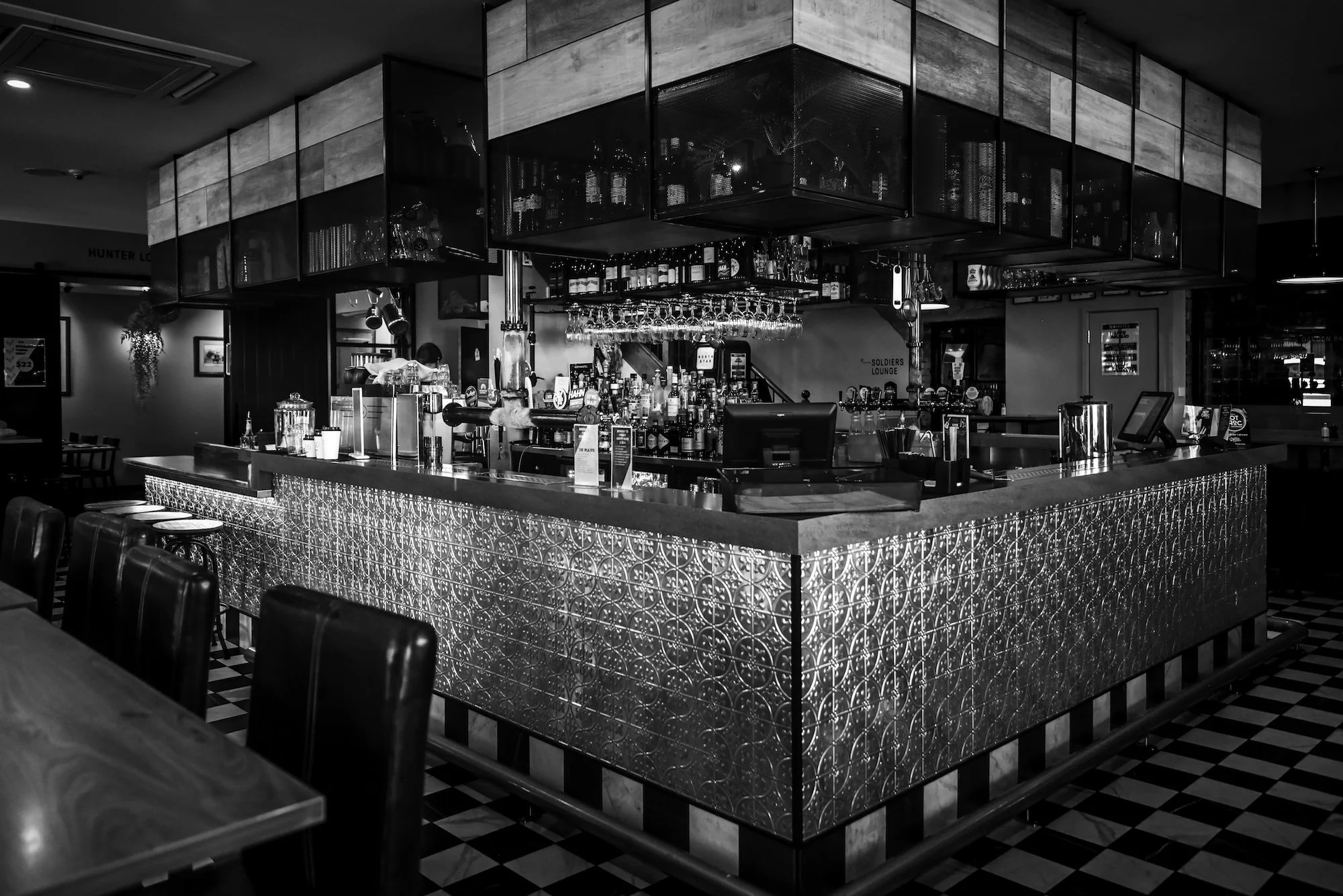 Black and white photo of a bar with a tiled bar counter and various bottles of alcohol behind it. Hanging glasses above the bar and chairs in front of the counter. Dark ceiling with lighting fixtures.