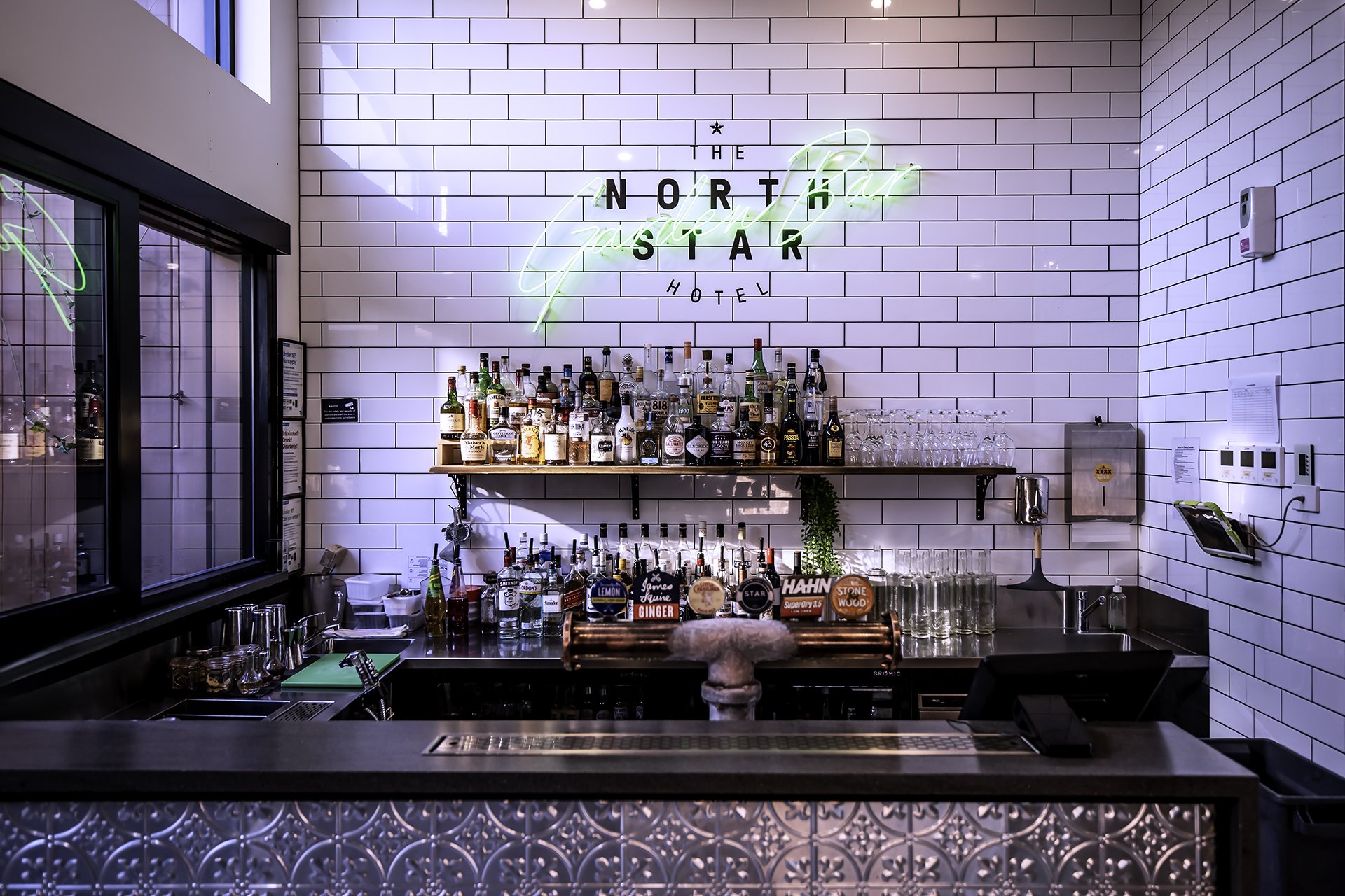 Interior of a bar with white tiled walls, a neon sign with the words 'The North Star Hotel', a collection of liquor bottles on a shelf, and various bar tools and glasses on the counter.