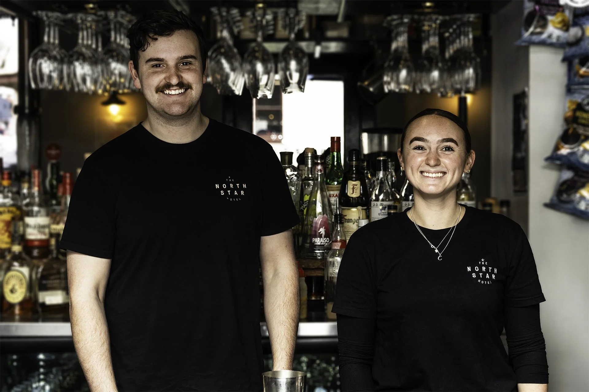 Two bartenders, one male and one female, standing behind a bar with liquor bottles and hanging glasses, smiling at the camera.