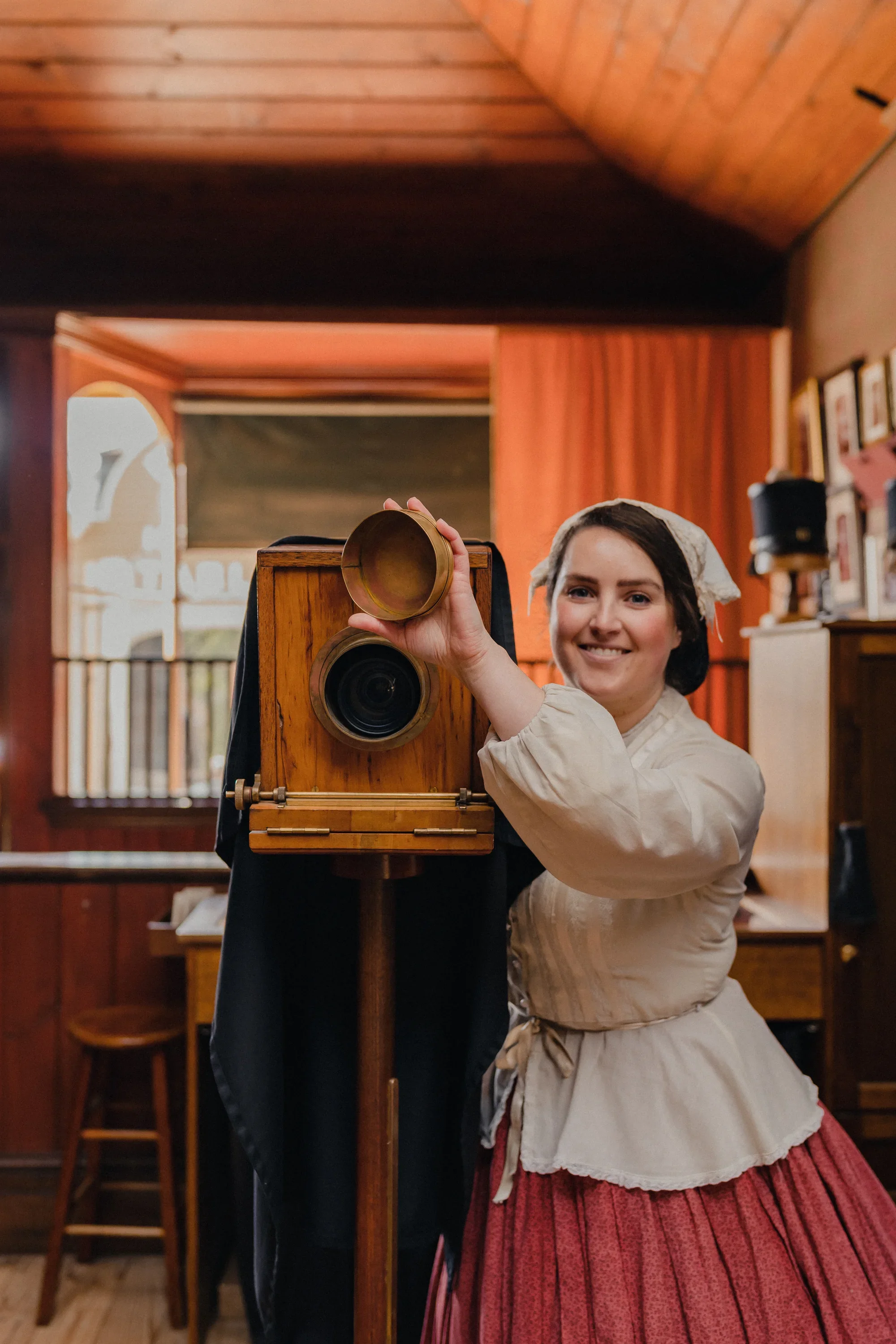 A woman dressed in historical clothing, smiling, holding a brass object next to an old wooden camera on a tripod inside a room with wooden paneled walls and a window.