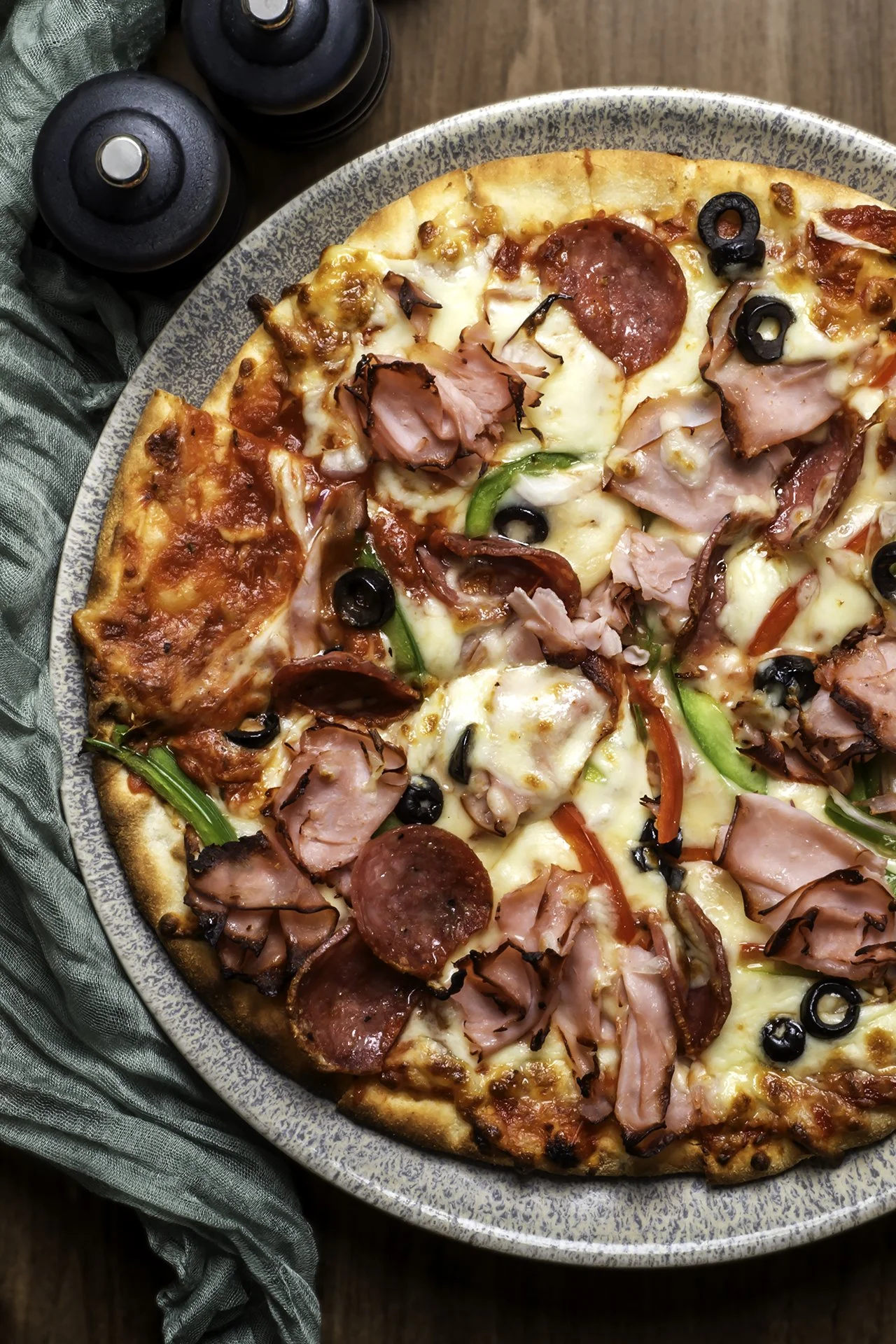 A pizza topped with pepperoni, ham, black olives, green peppers, and cheese on a grey ceramic plate.