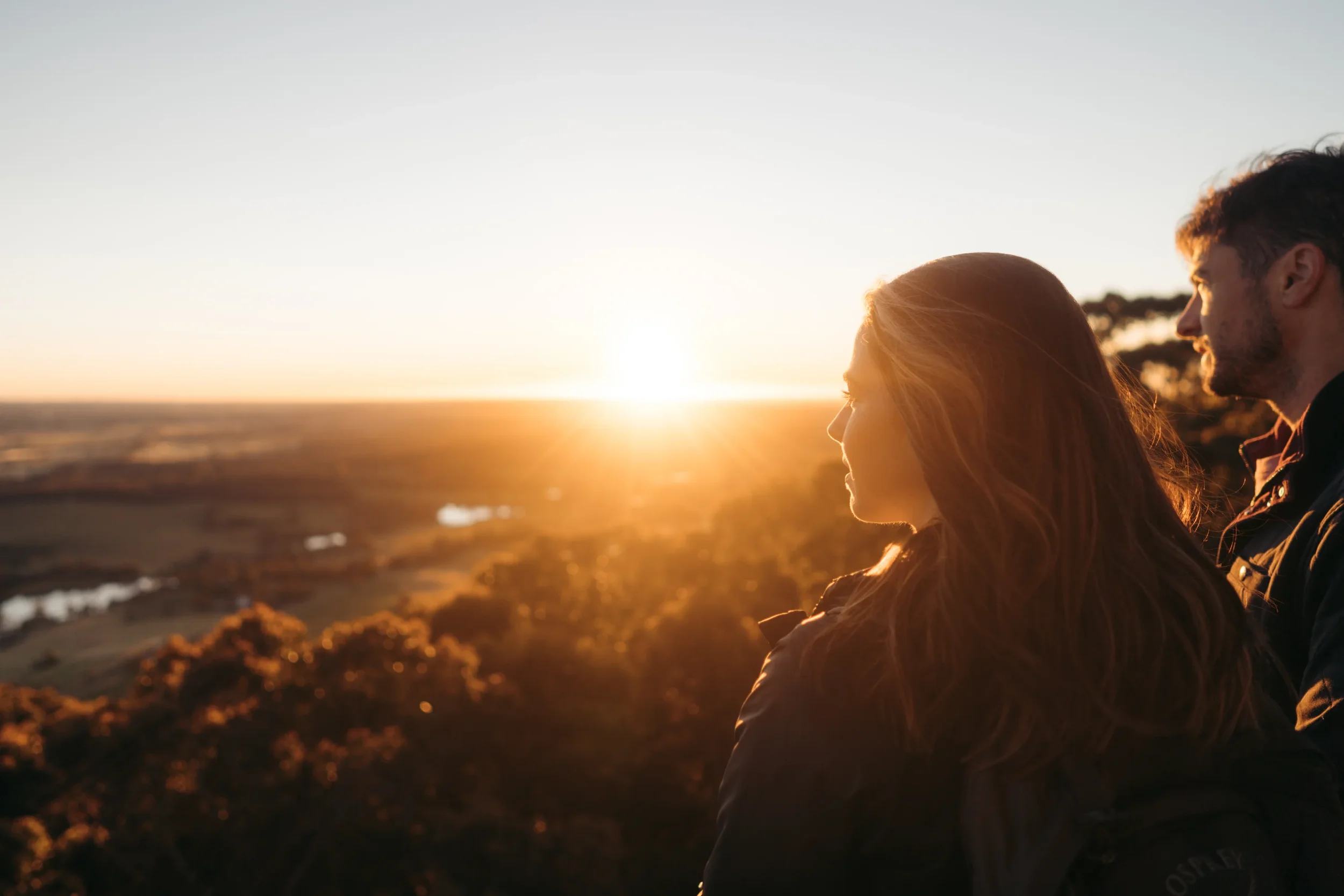 A man and a woman standing side by side outdoors during sunset, with warm sunlight illuminating their profiles, overlooking a landscape with trees and distant water.