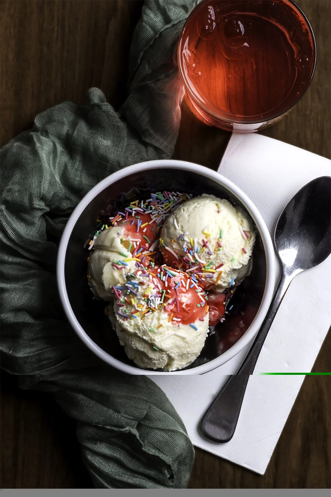 A bowl of vanilla ice cream topped with colorful sprinkles and pink syrup, a glass of red beverage with ice, a spoon, all placed on a white napkin on a wooden table.