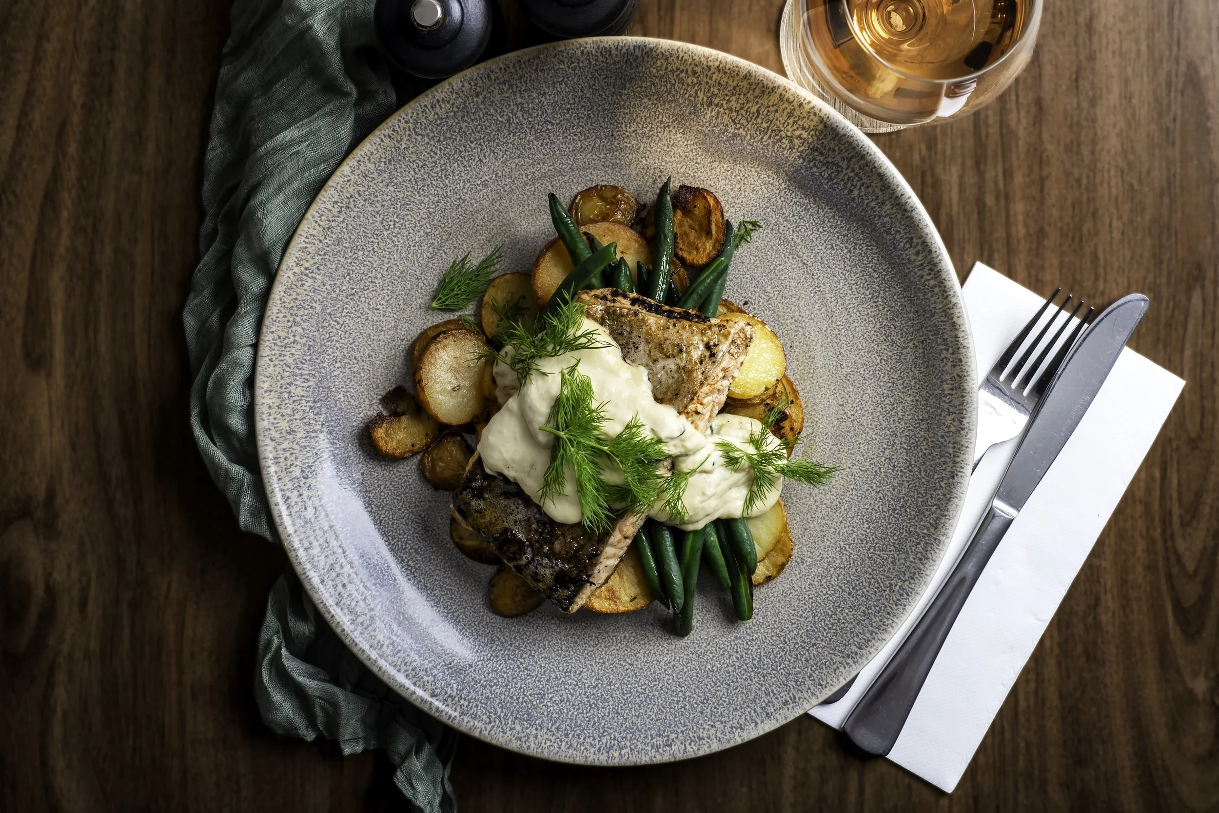 Plate with grilled salmon, roasted potatoes, green beans, and a dollop of cream sauce garnished with fresh herbs, on a wooden table with a glass of rosé wine, salt, and pepper shakers, and a fork and knife placed on a napkin.