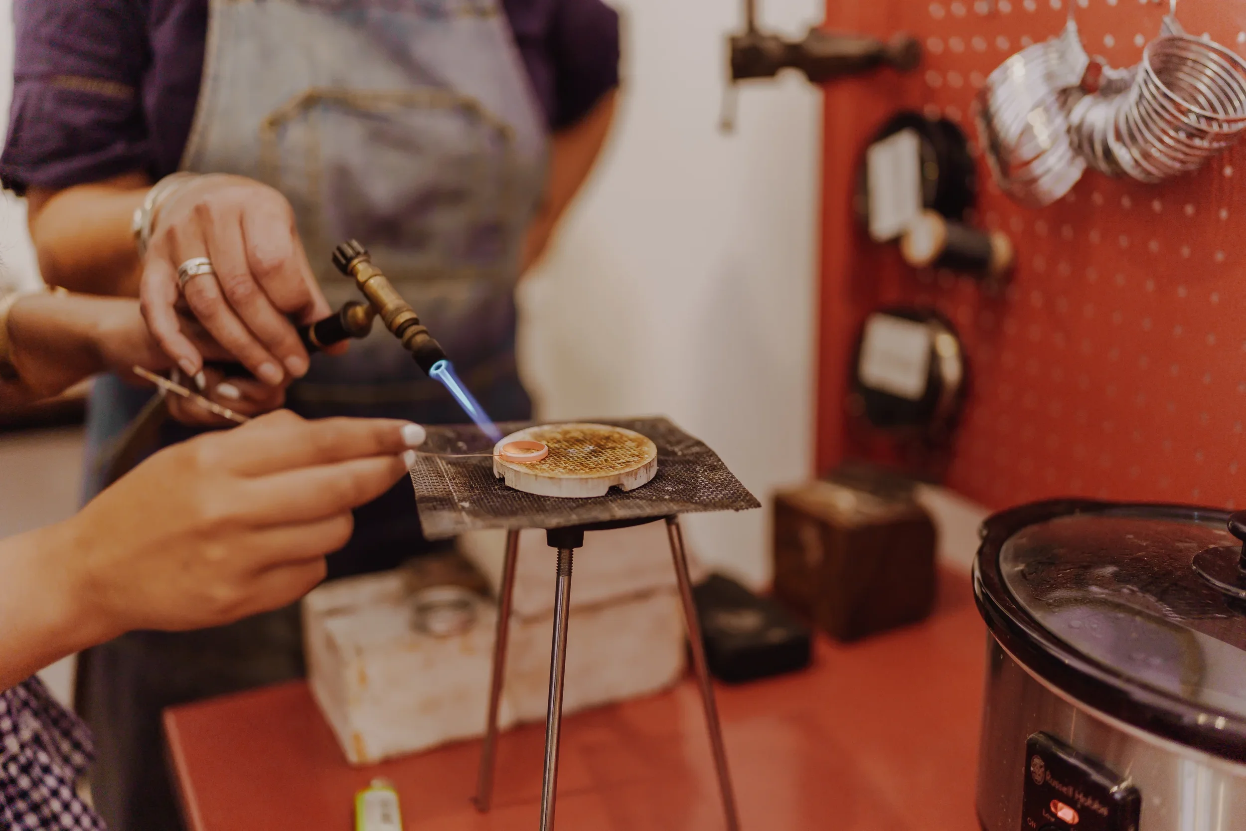 A person holding a torch or lighter near a small object on a metal stand, possibly performing a craft or scientific experiment in a workshop setting.