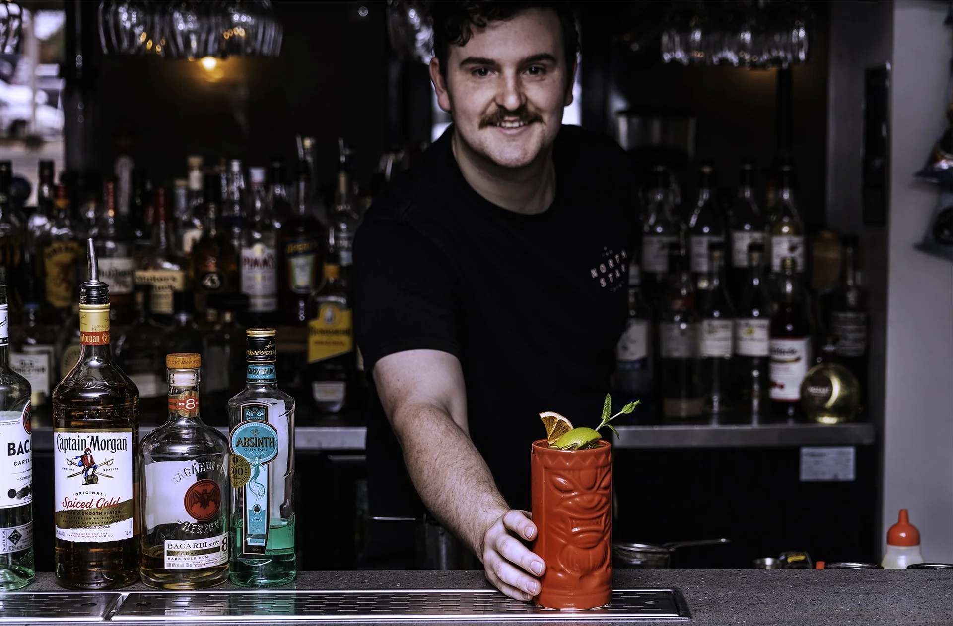 A bartender with curly dark hair and a mustache stands behind a bar, smiling at the camera. He is wearing a black shirt and preparing a colorful cocktail in a red tiki mug, garnished with fruit and mint. The bar has liquor bottles in the background a