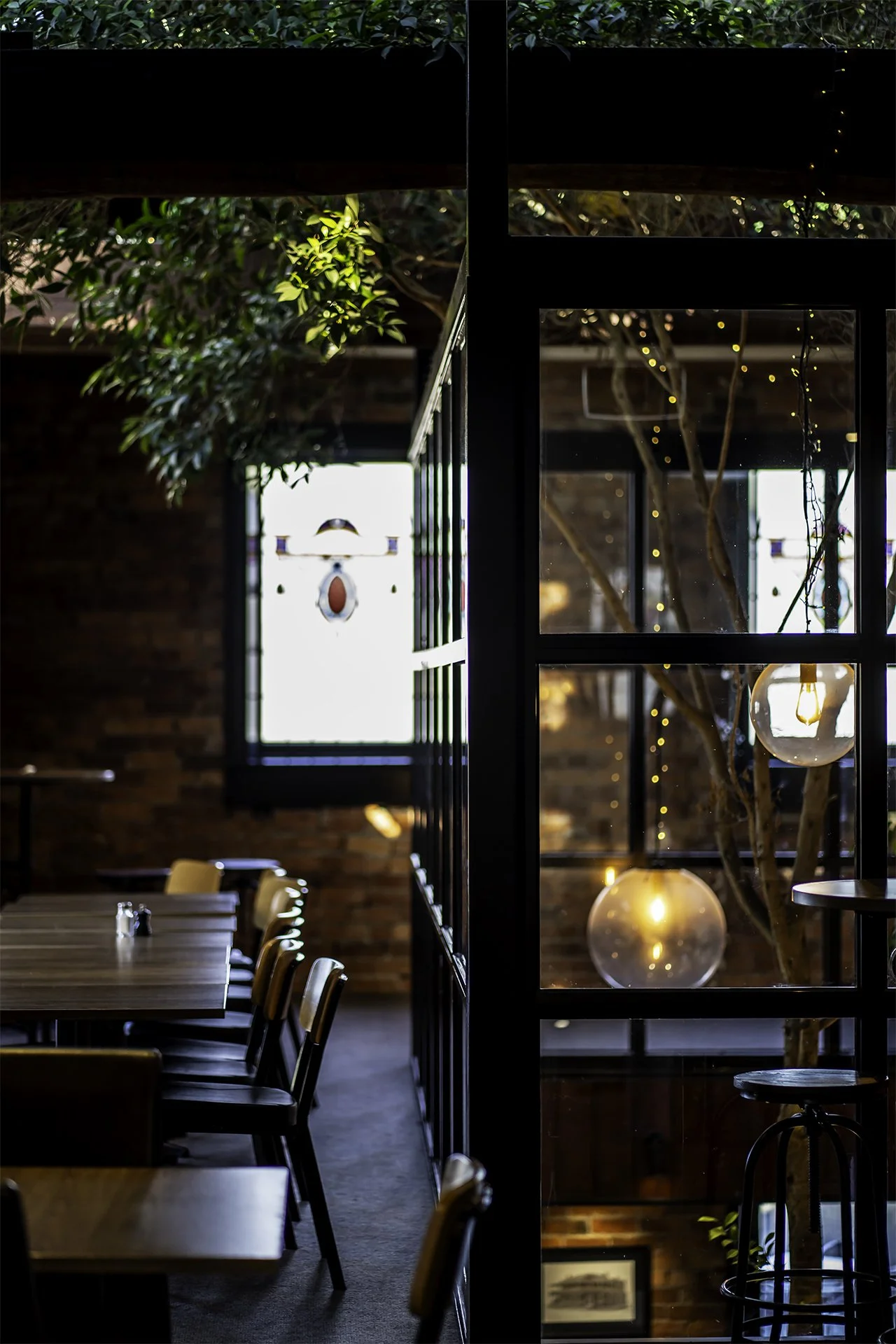 Interior of a modern restaurant or café with a wooden table, chairs, large windows, green plants, and warm lighting including hanging bulbs and string lights.