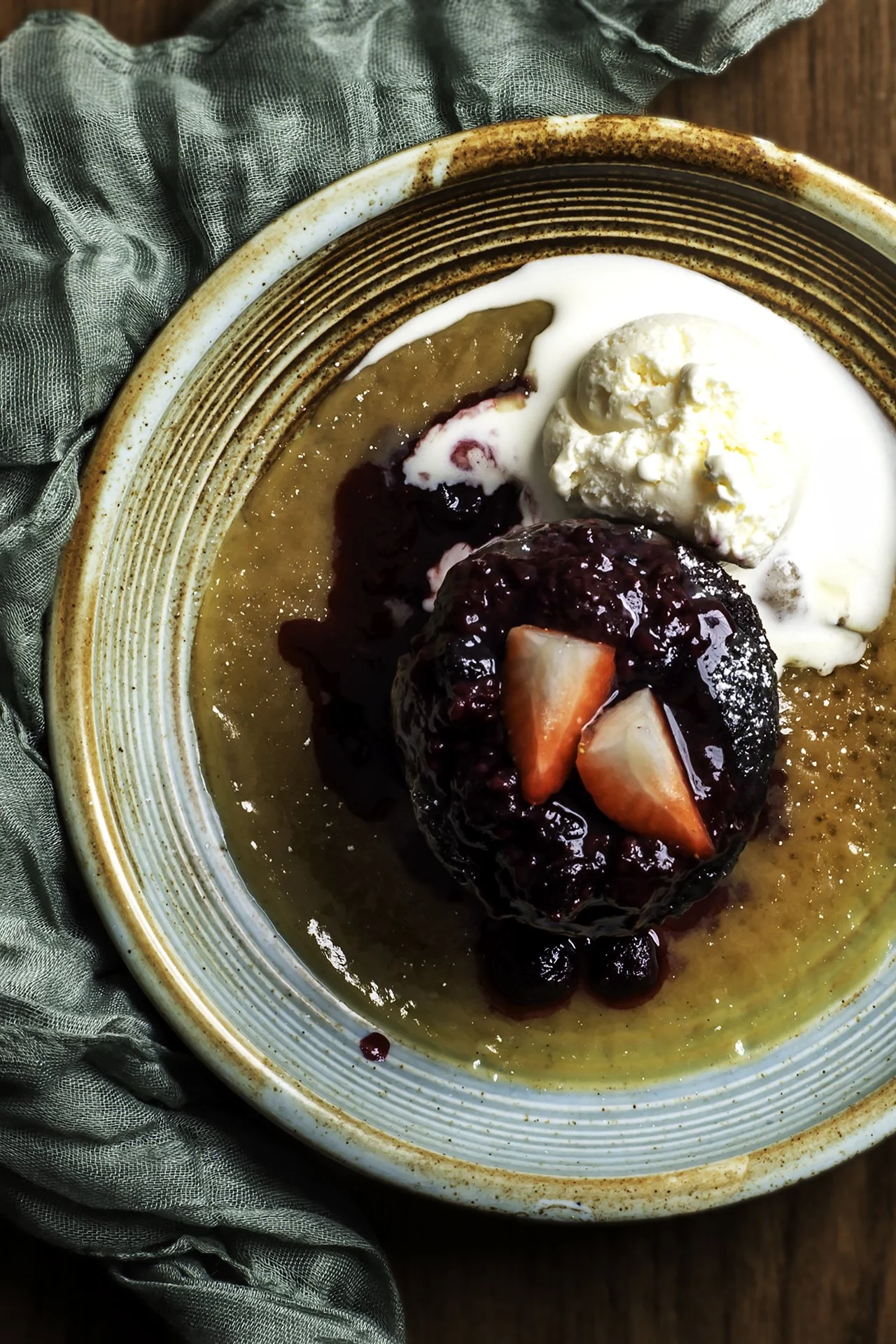 A bowl of caramelized pudding topped with berry compote, sliced strawberries, and a scoop of vanilla ice cream, surrounded by a green cloth on a wooden surface.