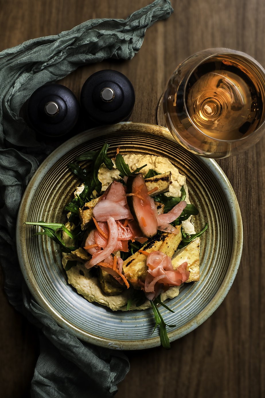 A plate of hummus topped with grilled vegetables and pickled onions, with a glass of rosé wine and condiments on a wooden table.