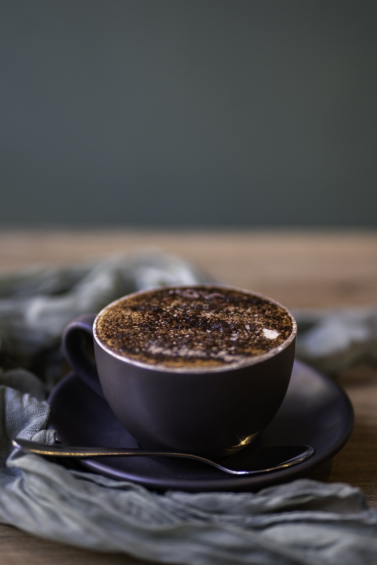 A cup of coffee with frothy top sprinkled with cocoa powder, placed on a black saucer with a silver spoon, on a cloth napkin with a wooden surface underneath.