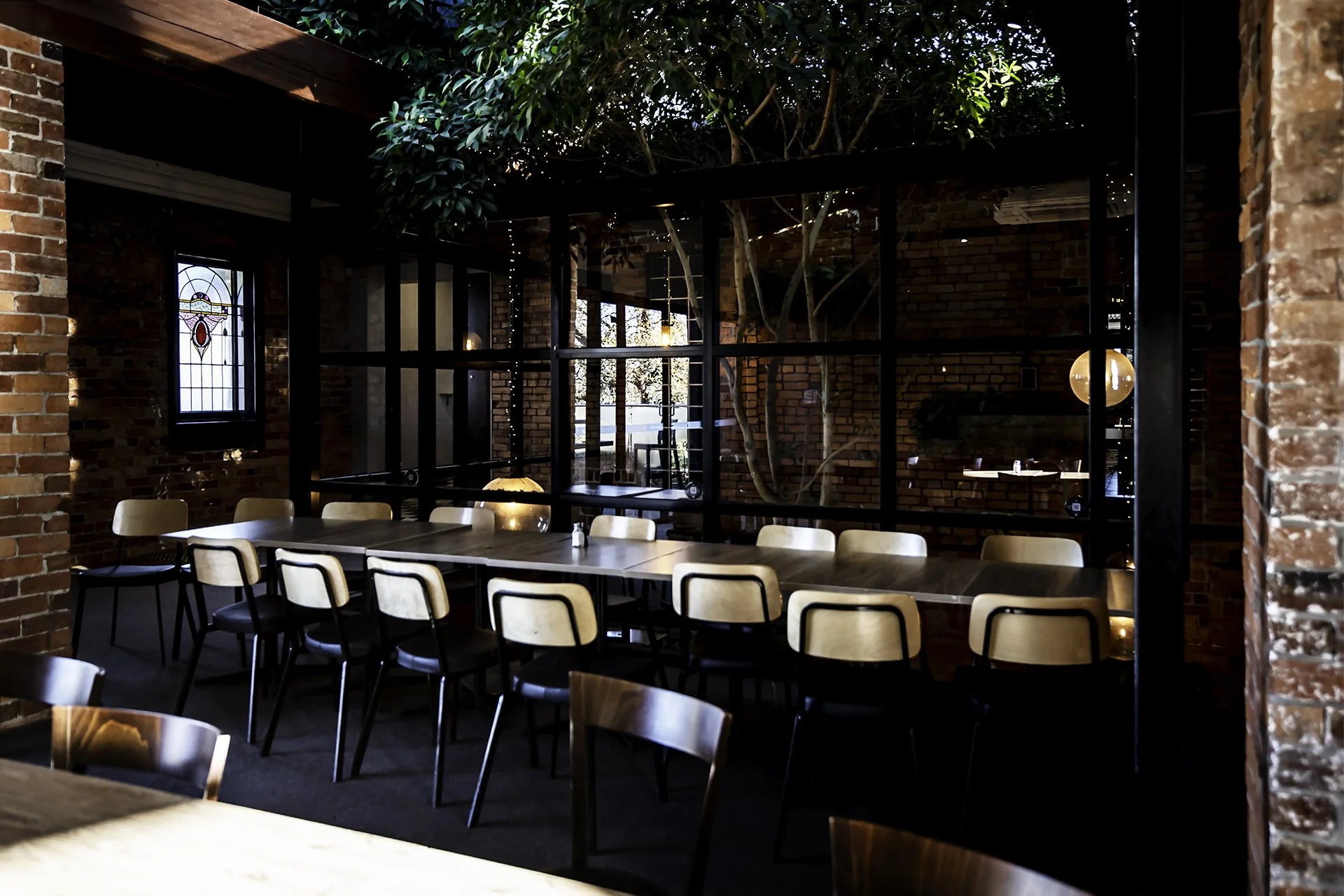 Interior of a restaurant with brick walls, a long wooden table surrounded by chairs, a large window, and a ceiling with a tree growing through it, creating a natural green canopy.