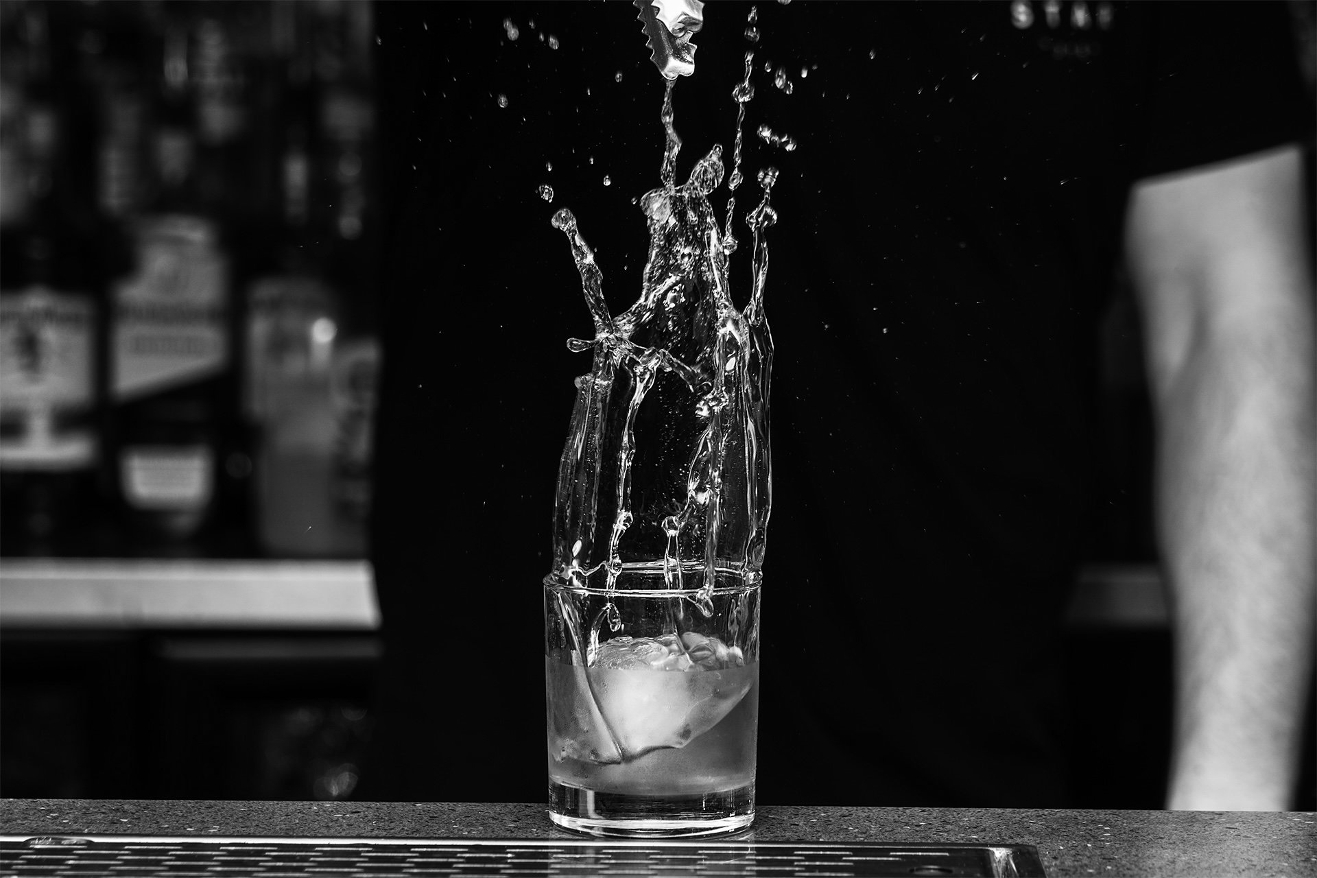 Close-up of water splashing out of a glass on a black countertop, with bottles blurred in the background.