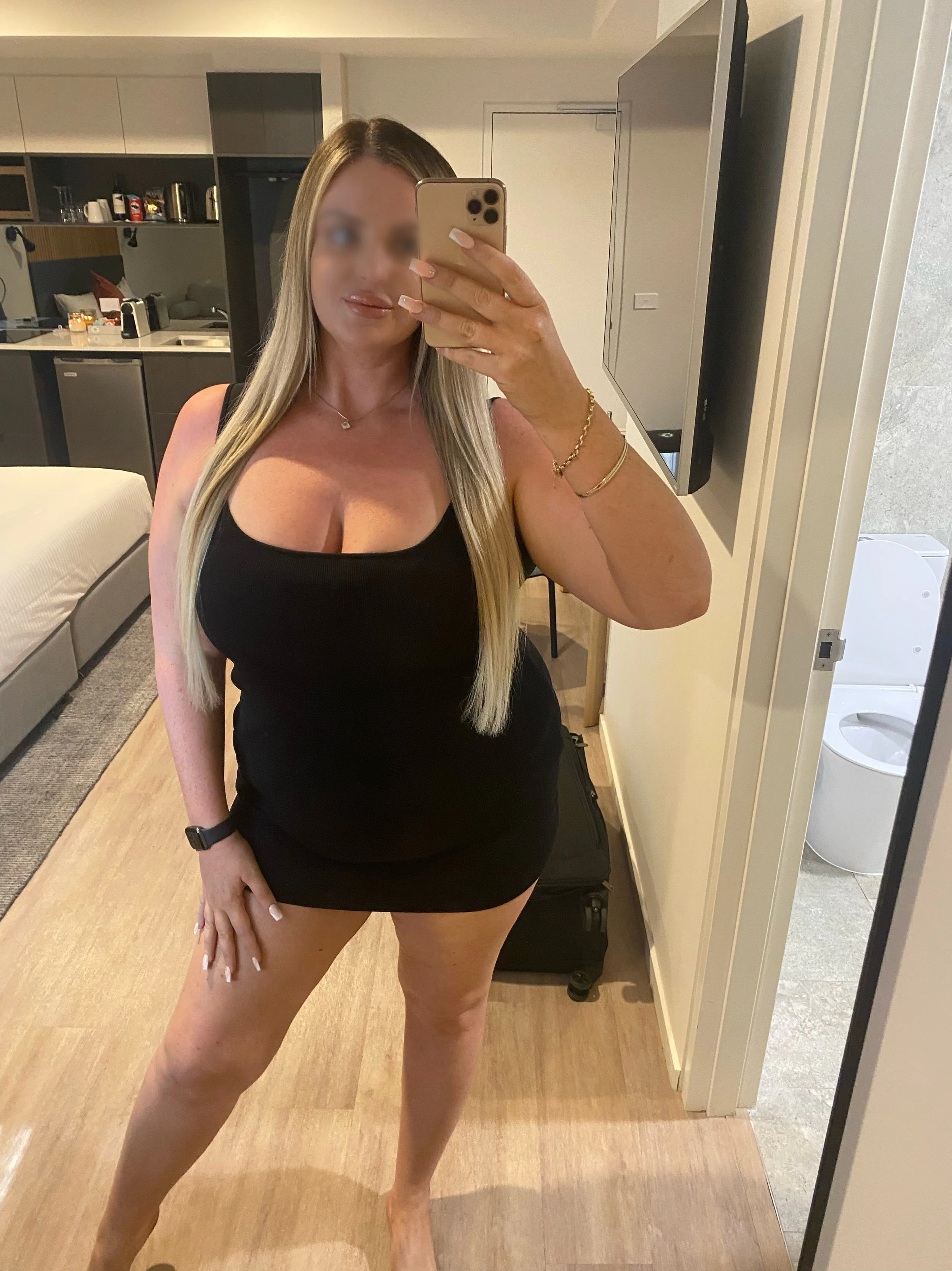 A woman taking a mirror selfie in a hotel room, wearing a black sleeveless dress, with long blonde hair, jewelry, and a watch, standing next to a black suitcase.