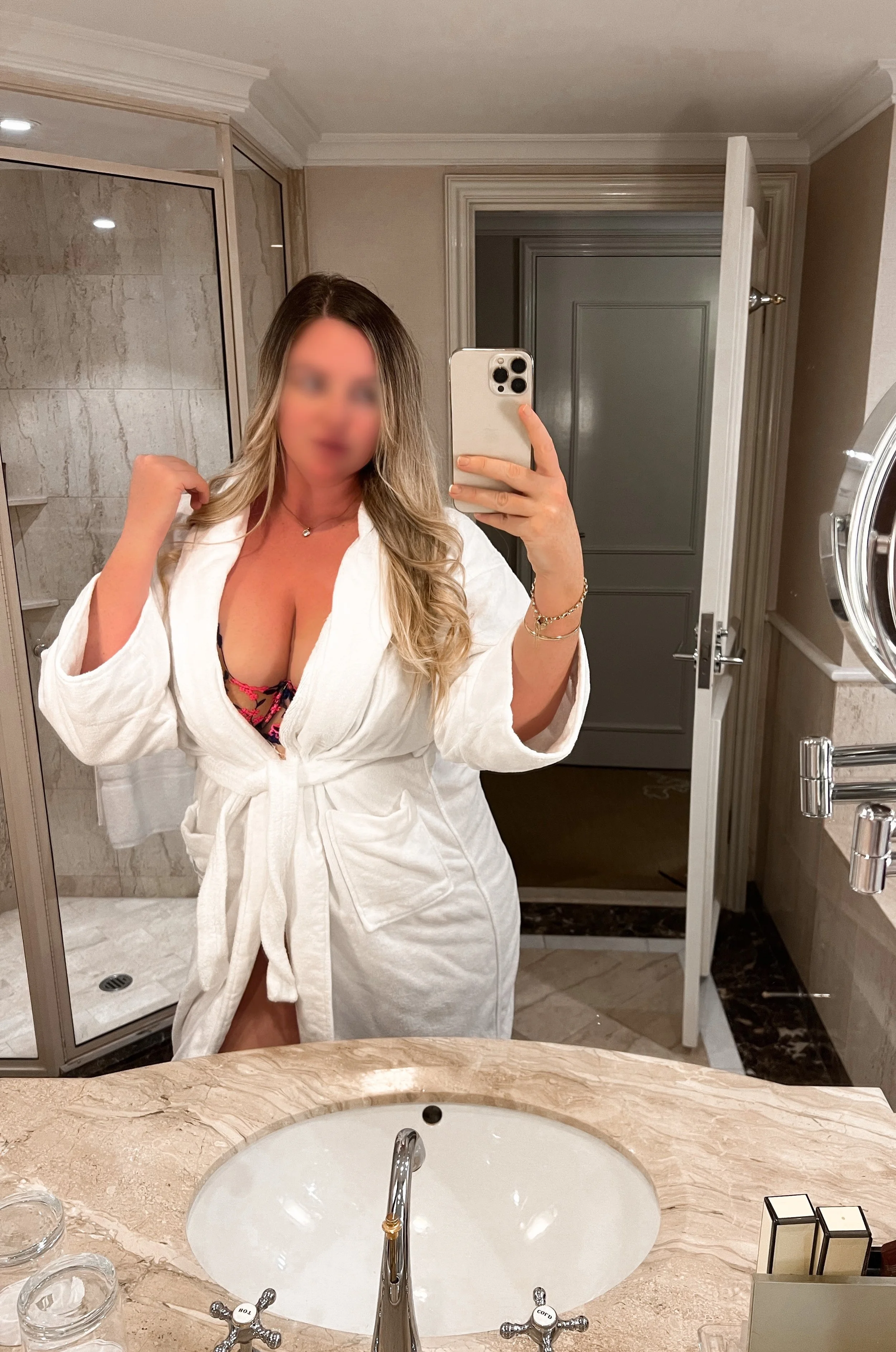 Woman taking a mirror selfie in a bathroom, wearing a white bathrobe with a black and pink lingerie top visible underneath.