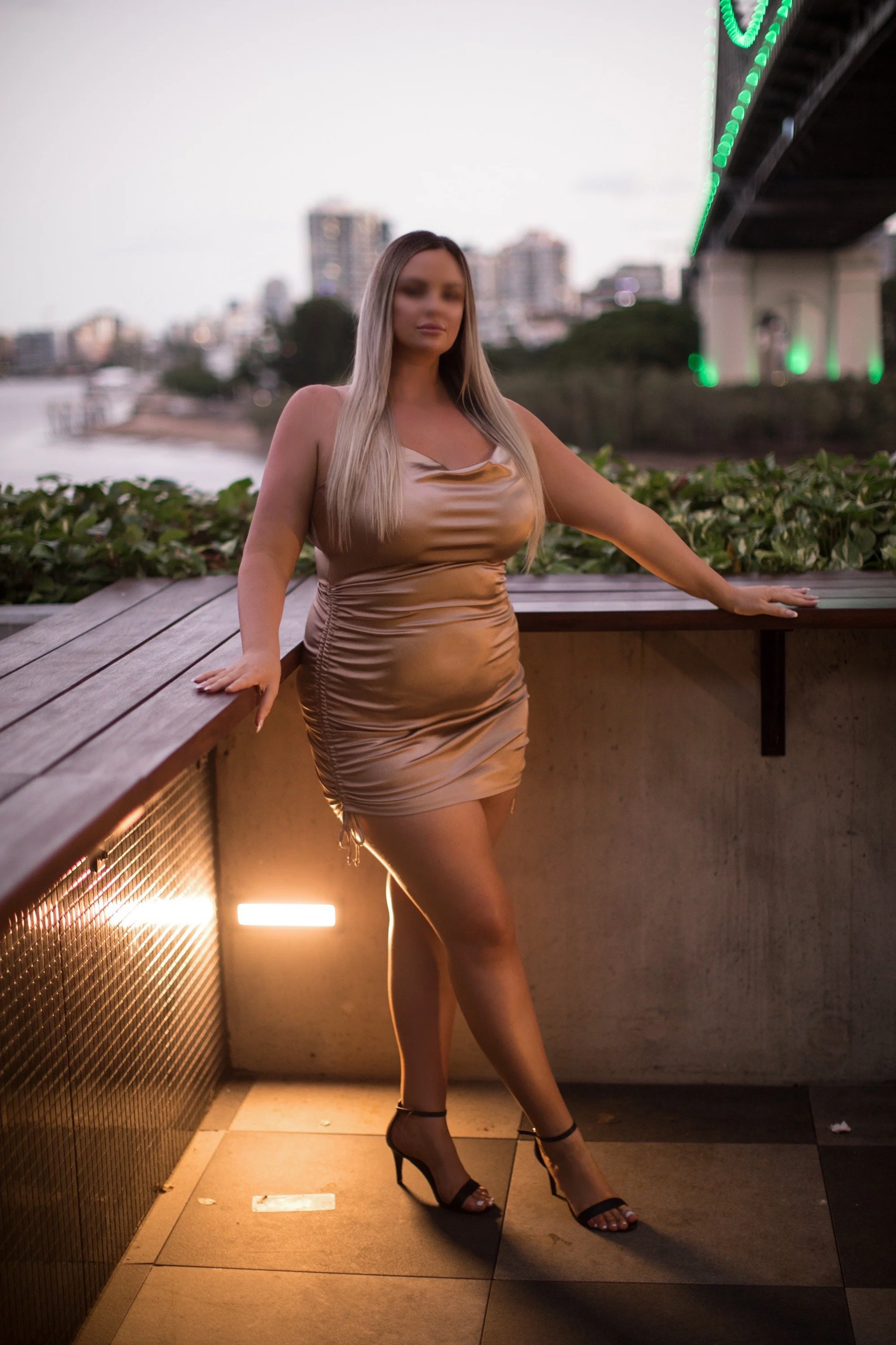 A woman wearing a shiny, beige satin dress and black high heels, standing outdoors near a railing with a cityscape and bridge in the background during dusk.