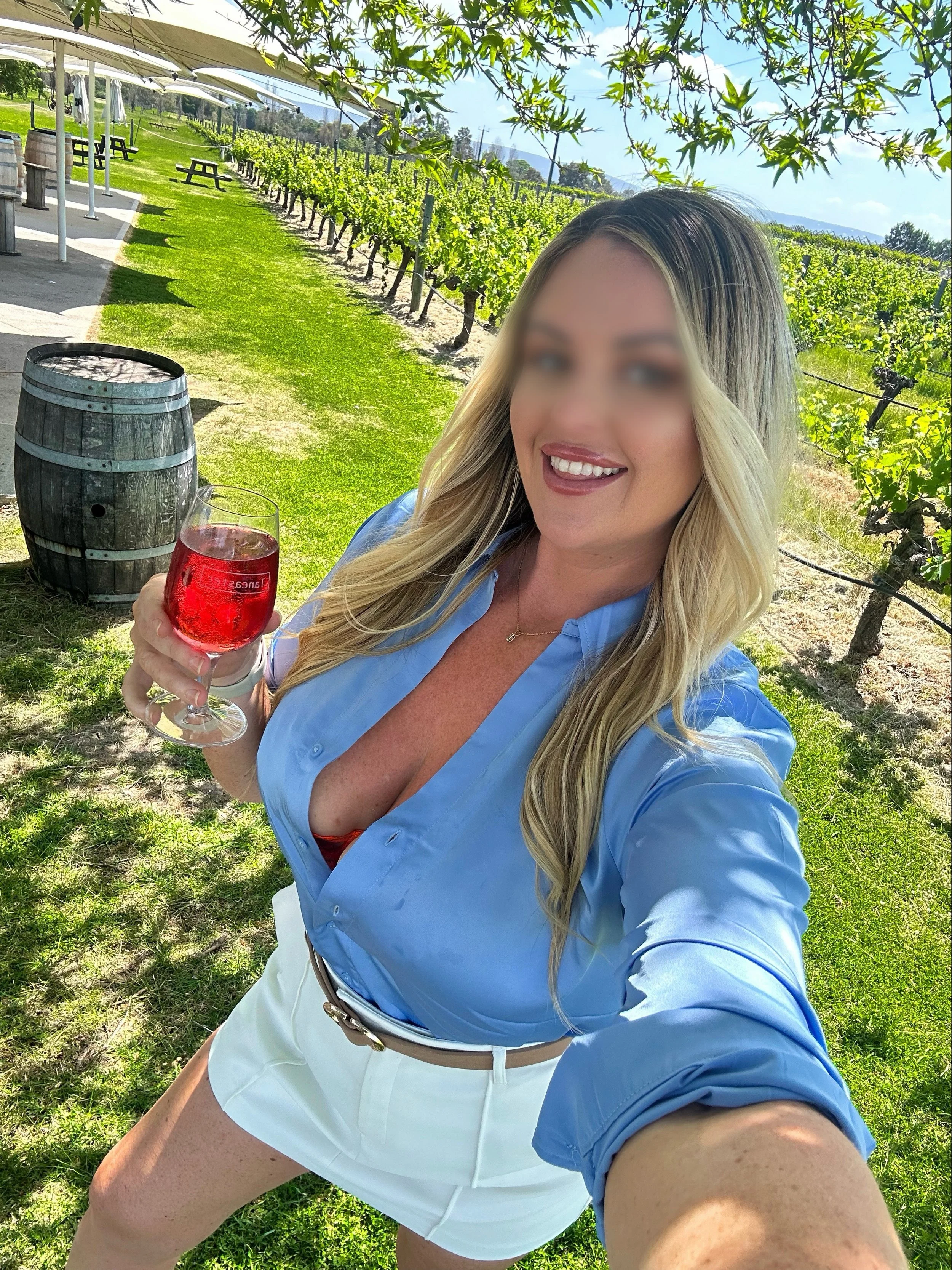 A woman with long blonde hair smiling and taking a selfie while holding a glass of red wine in a vineyard during daytime.