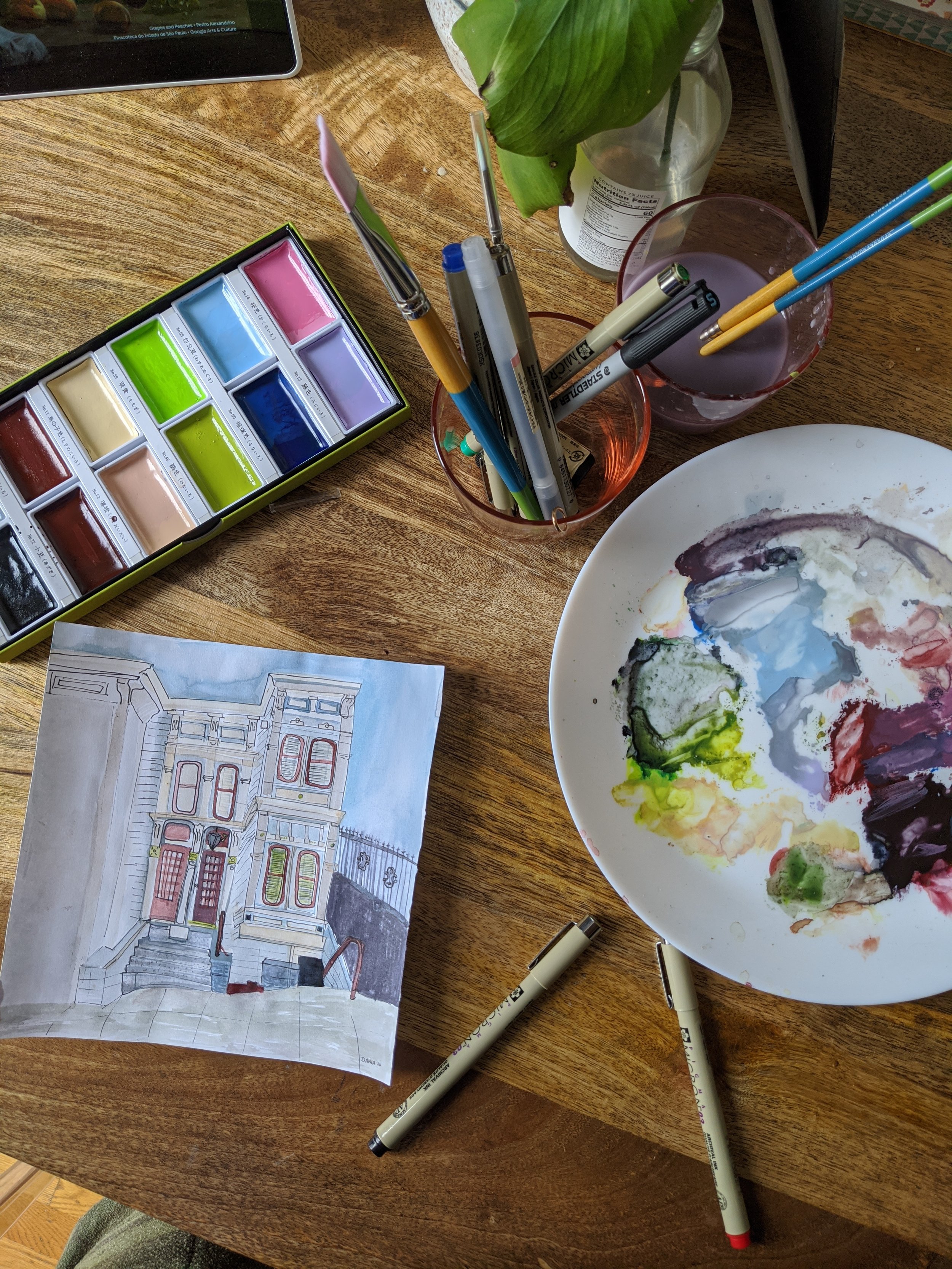 A watercolor painting of a historic building with steps, a small tree, and a wrought iron fence, on a wooden table surrounded by watercolor paints, markers, brushes, a palette with mixed colors, a cup of water, and a potted plant.