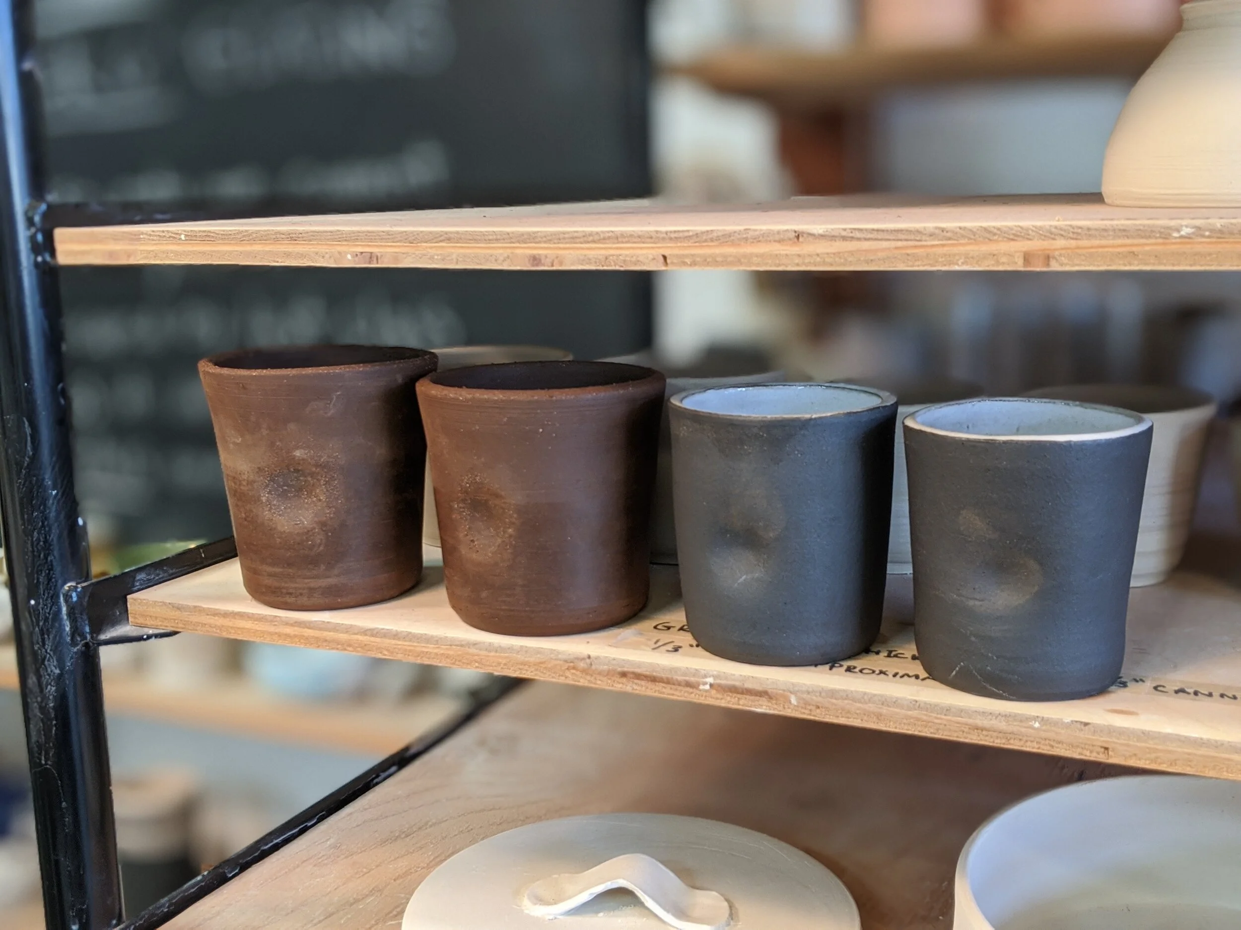 Four small ceramic cups, two brown and two dark gray, on a wooden shelf.