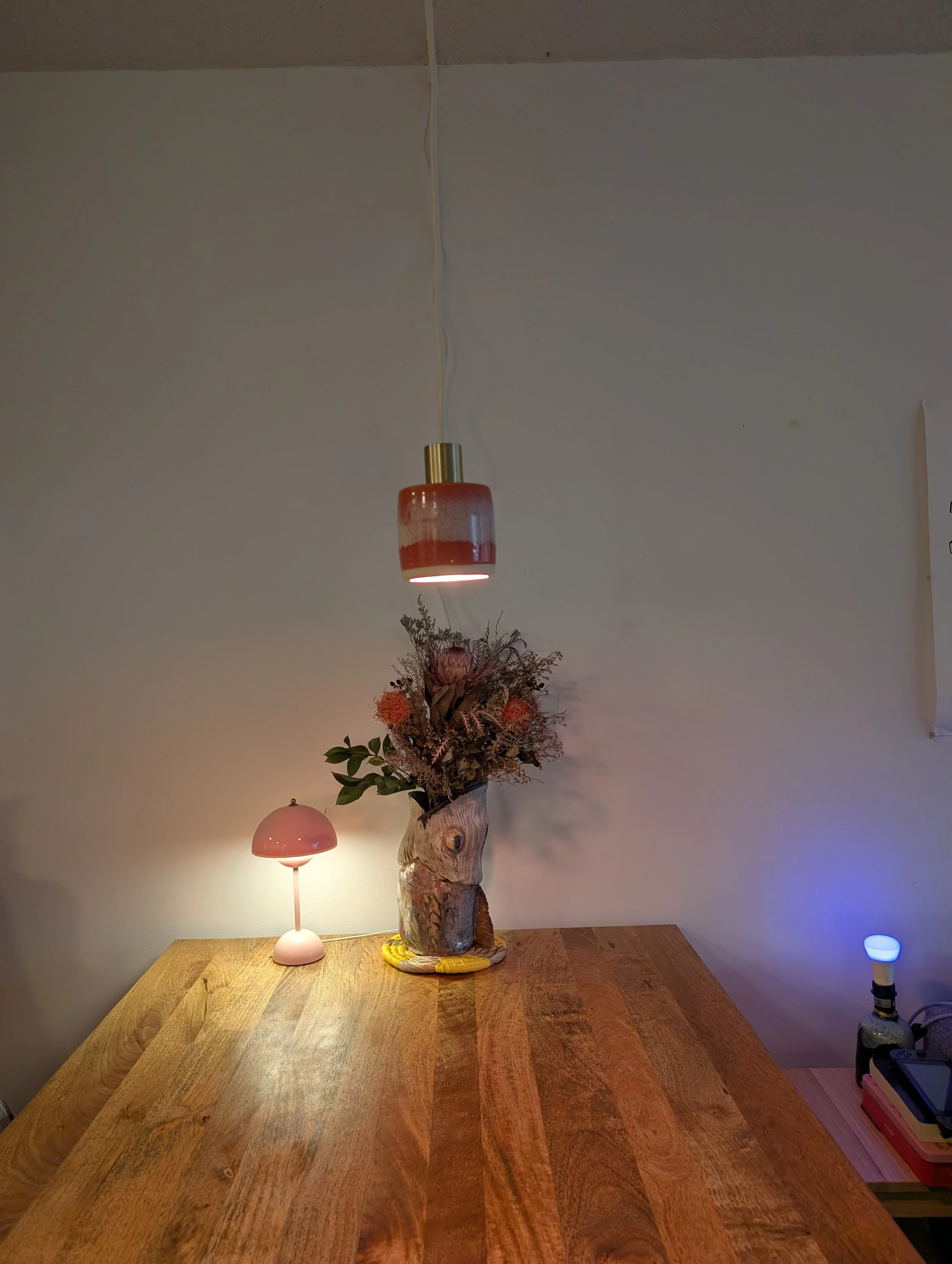 Decorative table with a carved wood tree trunk vase filled with dried flowers, a small pink lamp, a wall-mounted pink lamp, and a blue-lit object on a side table.