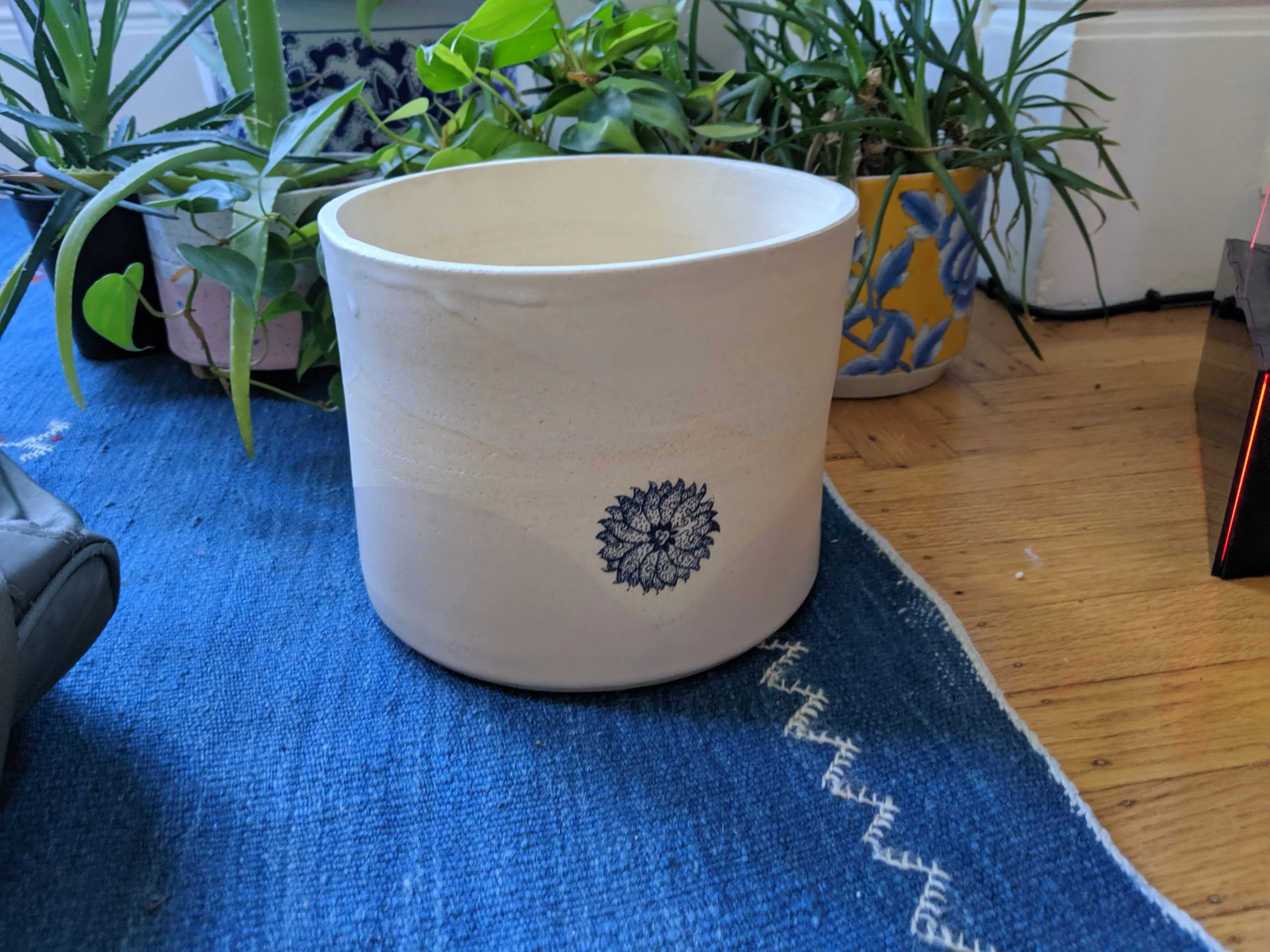 Empty white ceramic planter with a blue floral design, placed on a blue rug with a zigzag border, surrounded by green potted plants on a wooden floor.