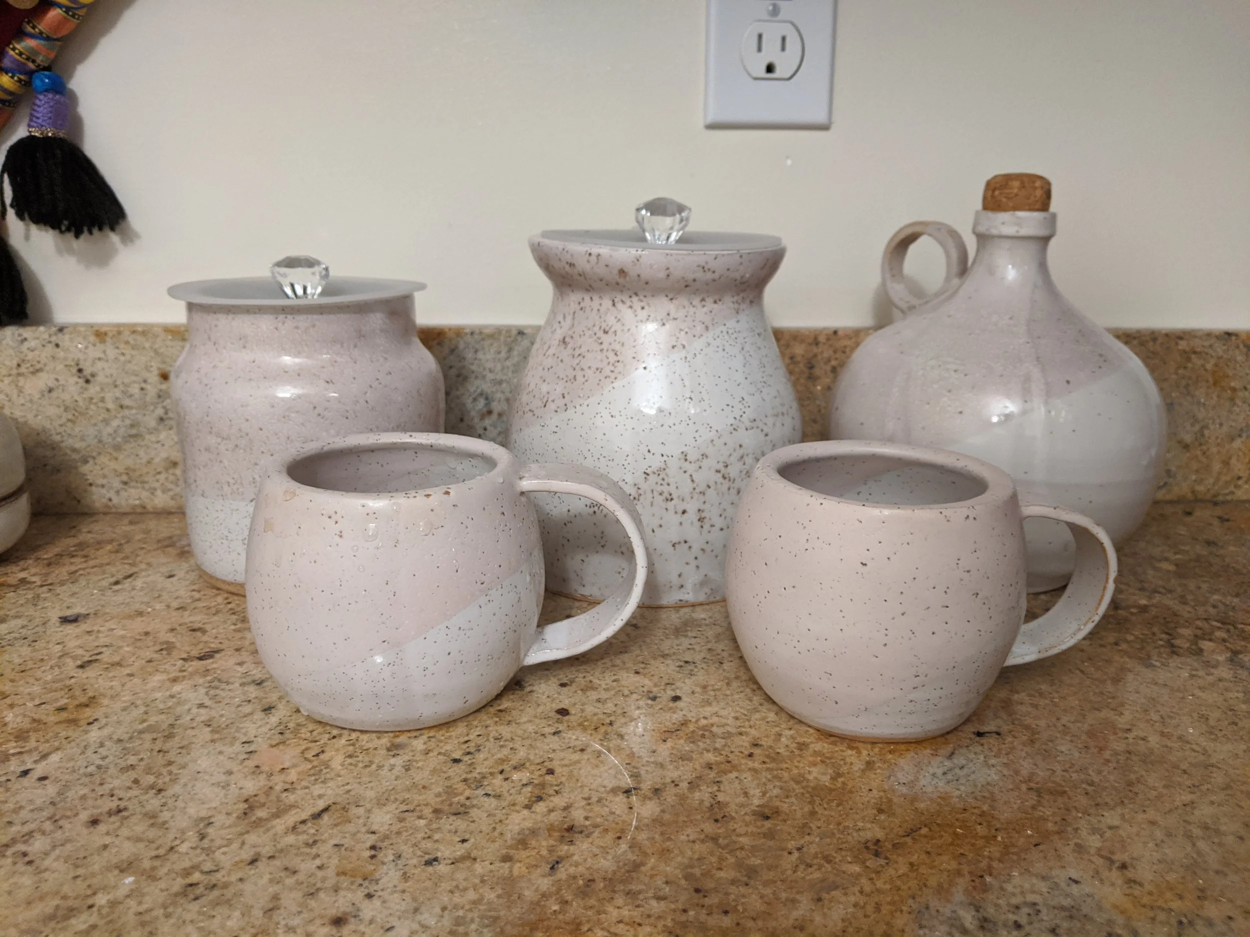 Six ceramic kitchen items on a granite countertop. There are two mugs with handles, two jars with lids that have decorative glass knobs, and a large bottle with a cork stopper.