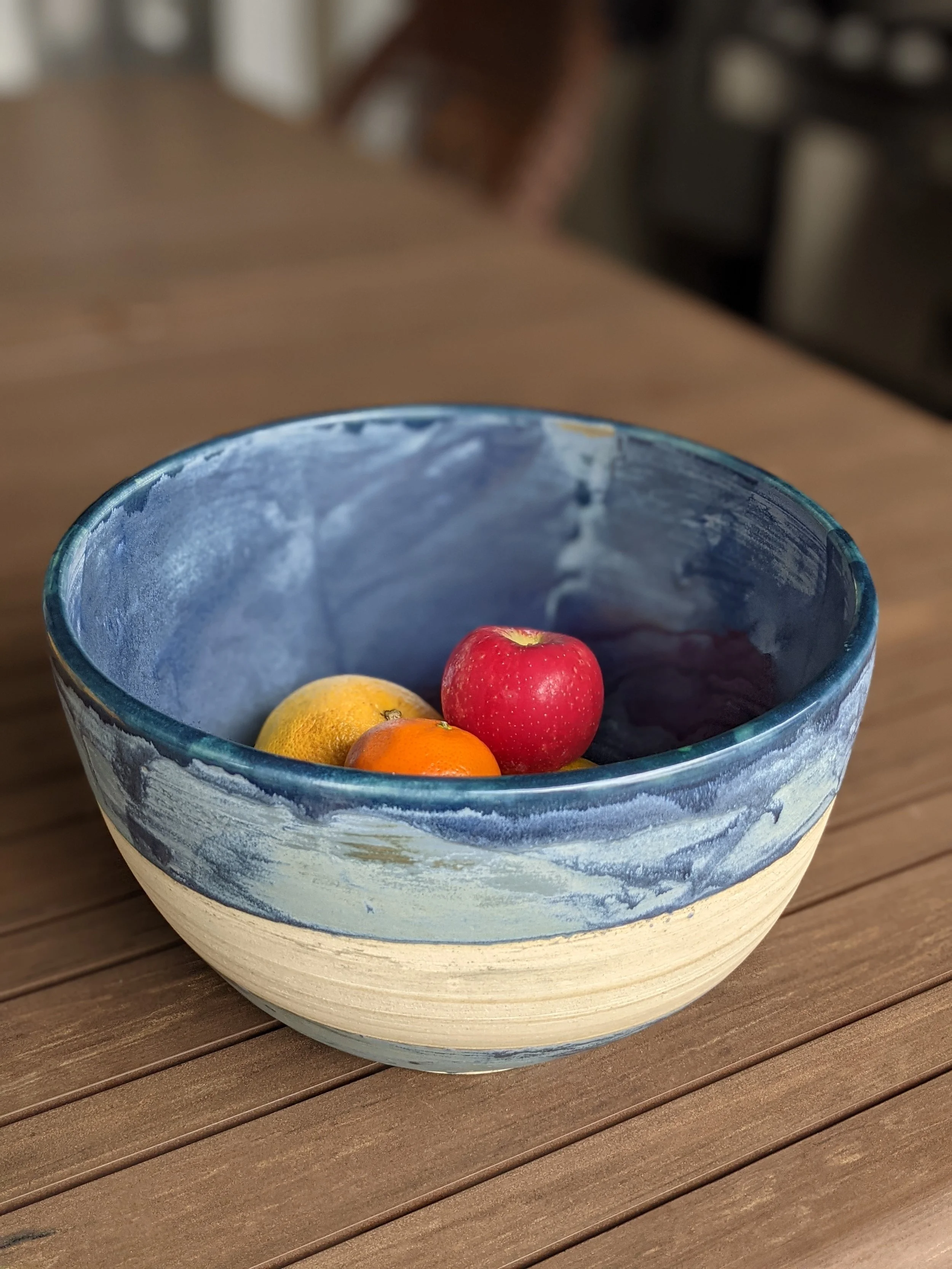A ceramic bowl with a blue and beige marbled design contains three small fruits: a red apple, a yellow lemon, and an orange or tangerine, placed on a wooden table.