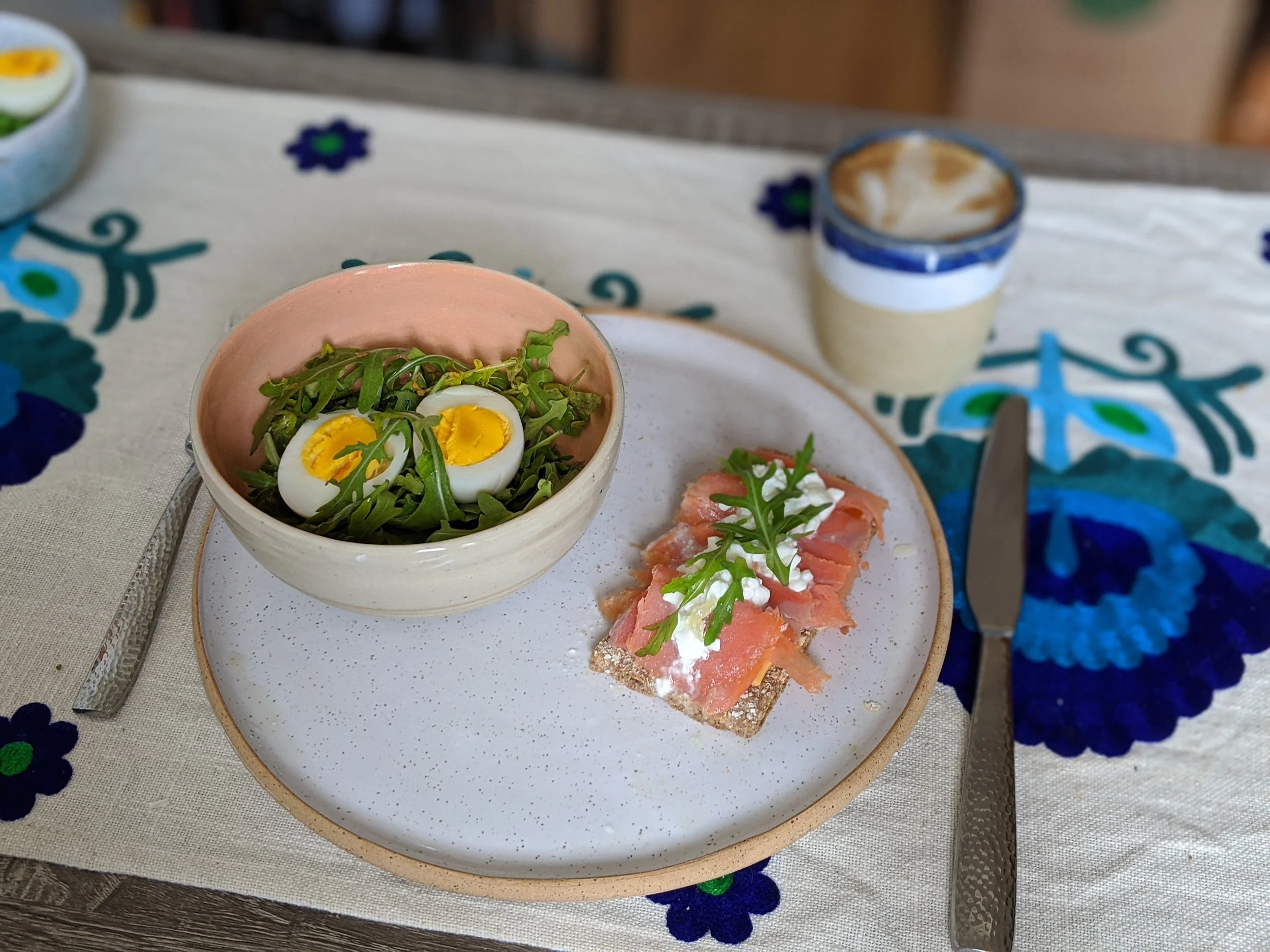 A plate with smoked salmon on bread topped with cream cheese and arugula, a bowl of salad with greens and sliced boiled eggs, a knife, and a cup of coffee on a floral tablecloth.