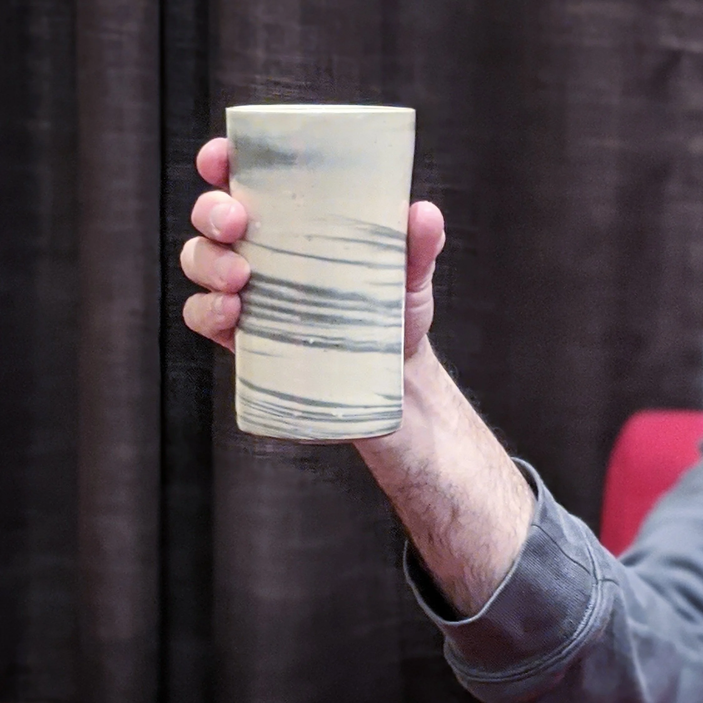 A hand holding a tall ceramic mug with a cream and gray marbled pattern, against a dark background.
