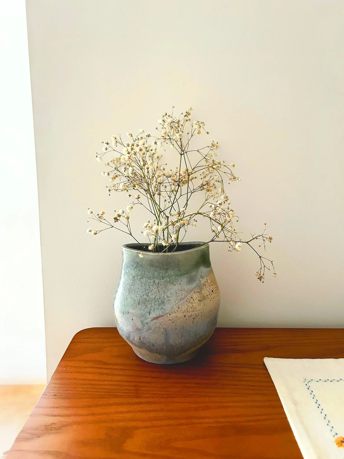 A ceramic vase with a glaze of green, white, and beige, containing dried white flowers with small blossoms, placed on a wooden table next to a white cloth with blue embroidery.