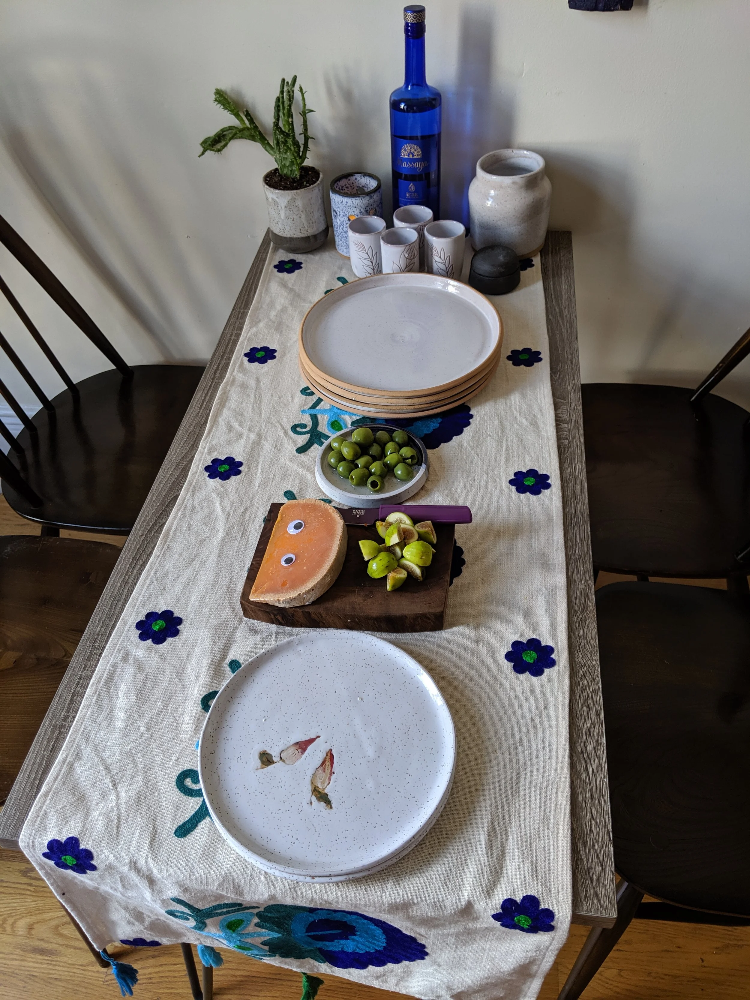 Dining table with plates, a cheese wedge, olives, a knife, and a table runner with blue and green floral embroidery, surrounded by three chairs.