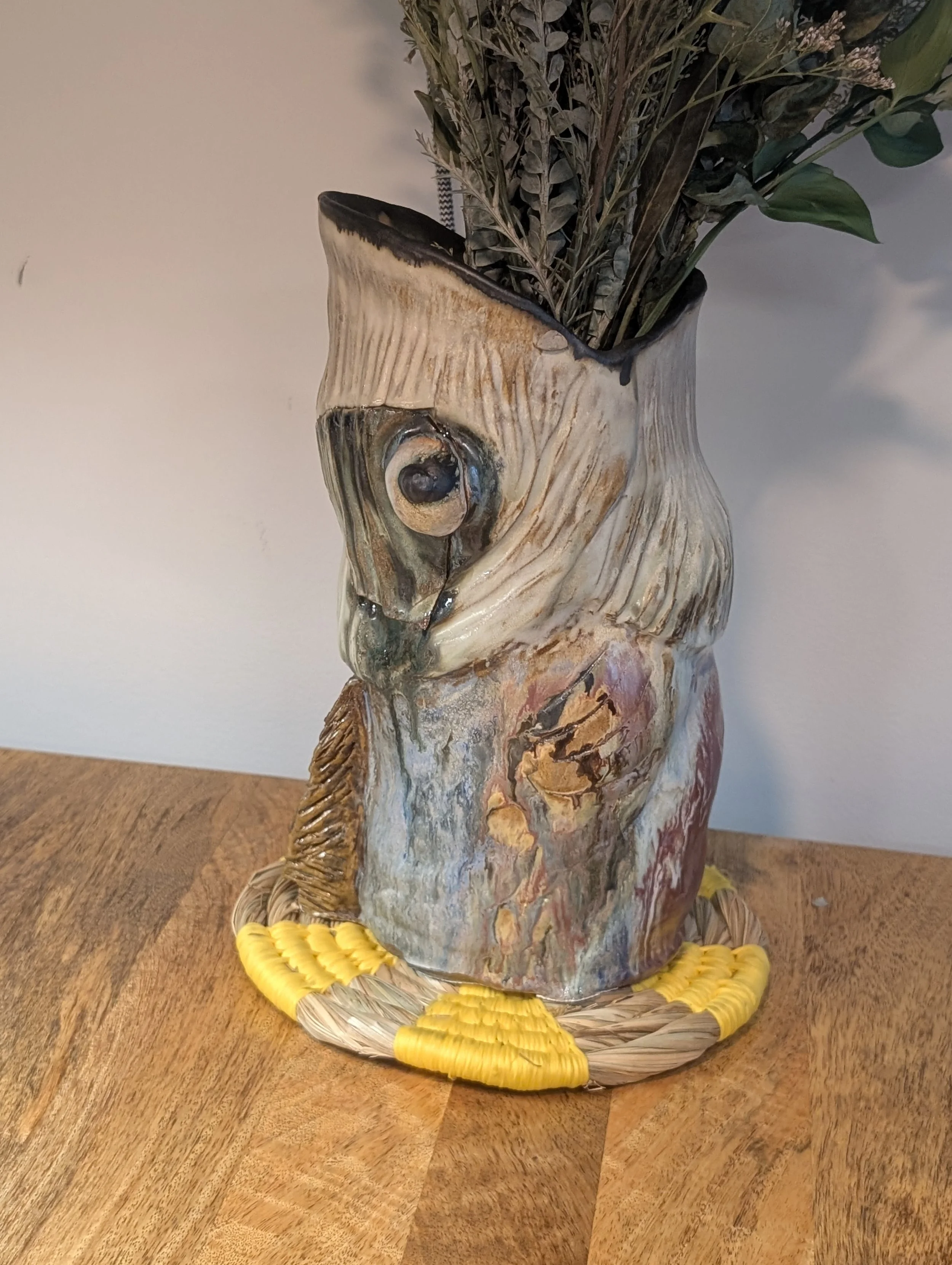A ceramic owl-shaped vase with a textured surface, filled with dried plants and leaves, sitting on a wooden table with a yellow decorative piece around its base.