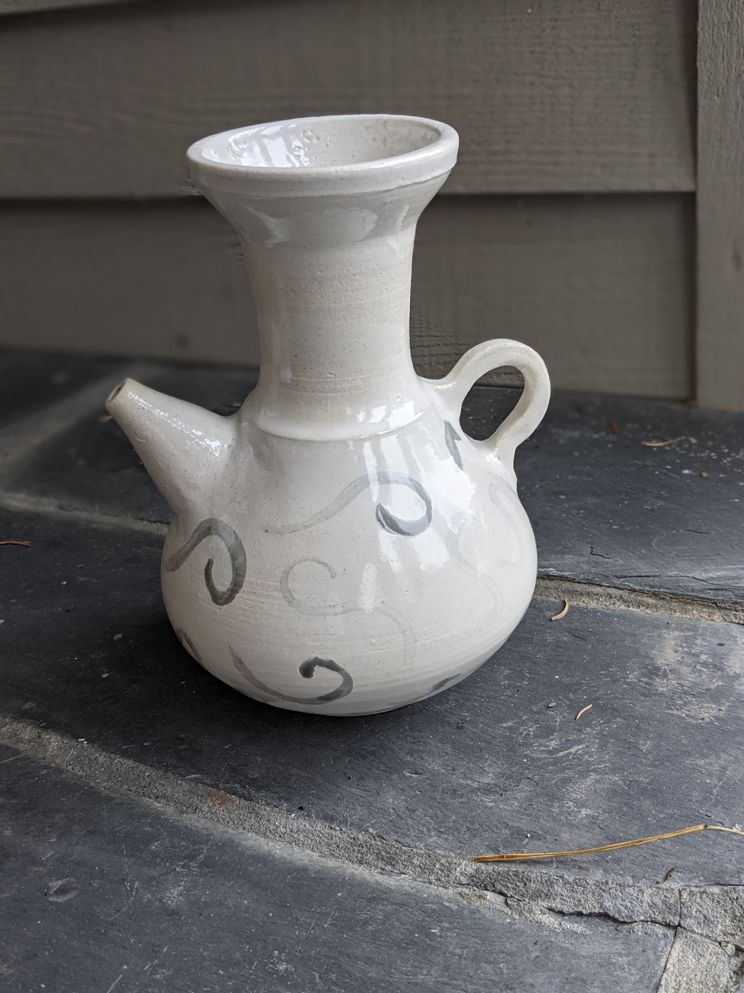 A ceramic teapot with a rounded body, a spout, a handle, and an elevated funnel-shaped top, decorated with gray swirly patterns, placed on a dark stone surface with a wooden wall background.