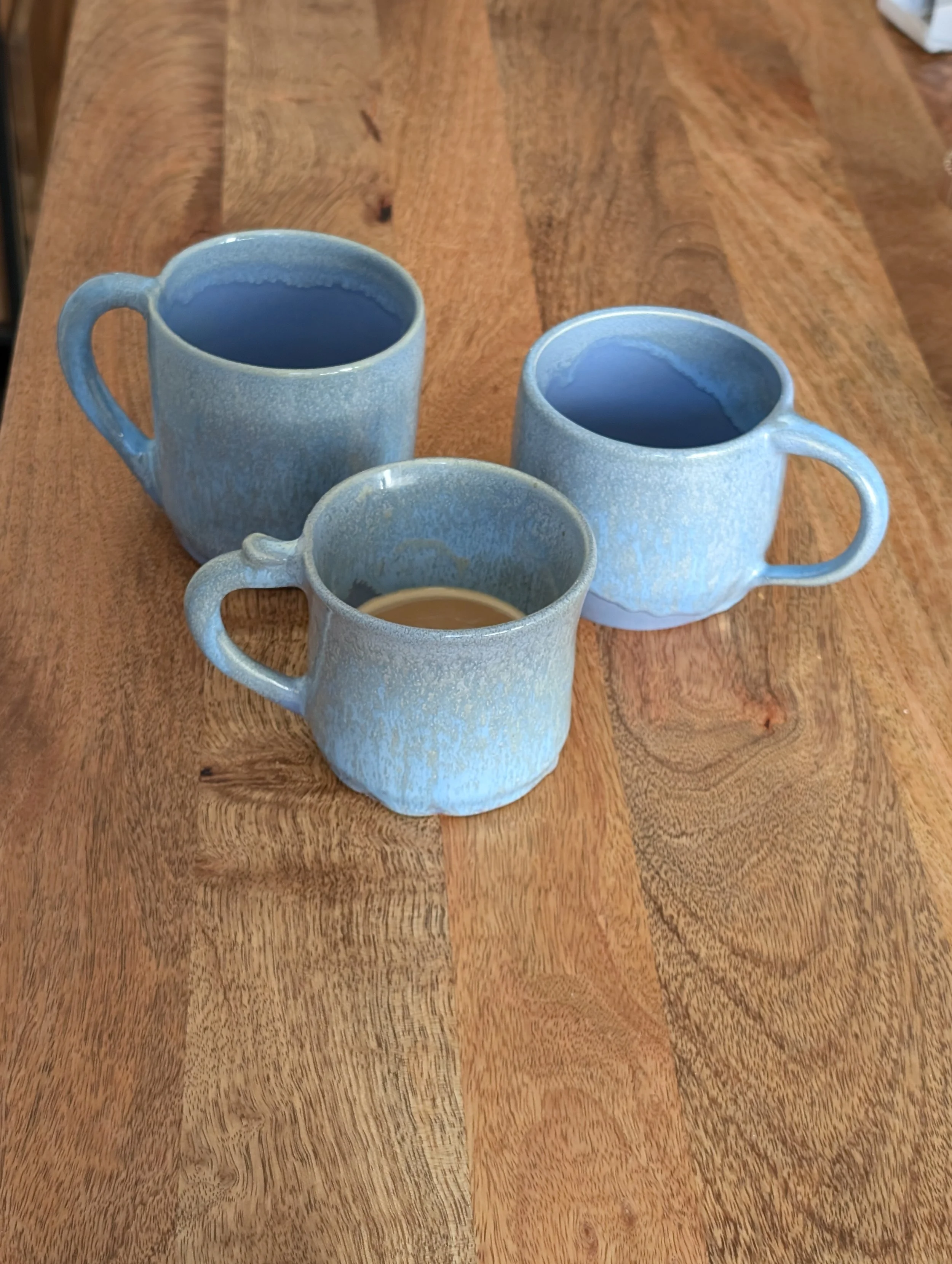 Three ceramic coffee mugs with handles, two larger ones with blue glaze and one smaller with a lighter glaze, placed on a wooden table.