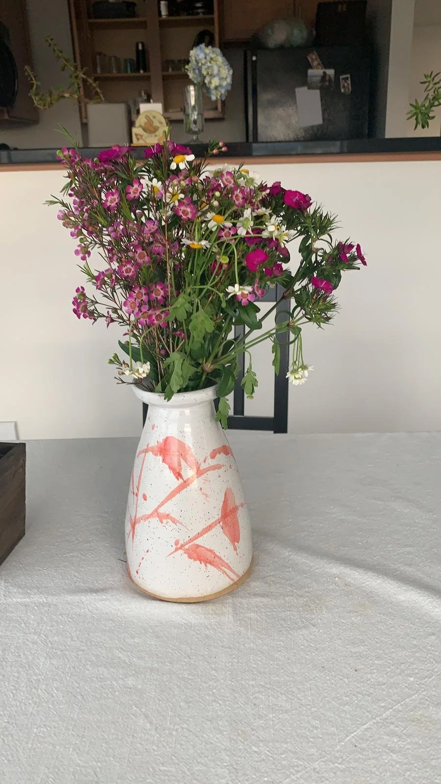 A white ceramic vase with pink and red abstract floral patterns, filled with assorted pink, white, and purple wildflowers, placed on a table with a white tablecloth.