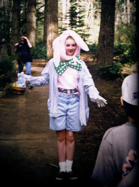 A person dressed in a bunny costume with a white hat and green polka-dot bowtie, standing on a wooded trail, holding a basket, with children and adults nearby.
