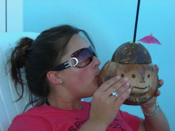 A woman wearing sunglasses and a pink shirt is drinking from a coconut with a carved face and a pink cocktail umbrella.