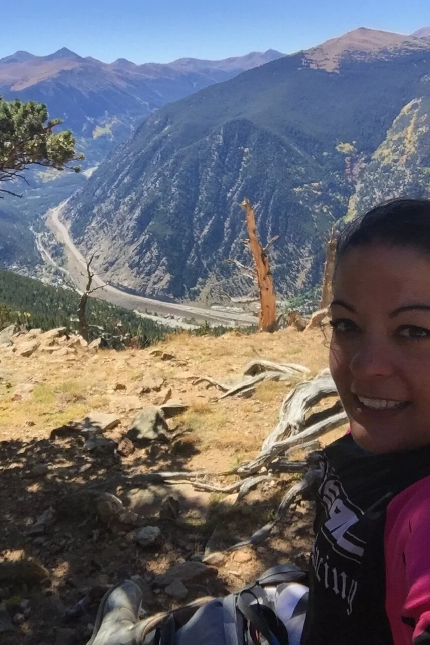 A woman taking a selfie in a mountainous landscape with a valley, trees, and a winding road in the background.