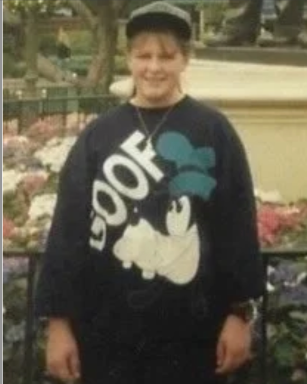 Person wearing a black hoodie with a Disney Mickey Mouse design and the word 'Joy' on it, standing outdoors in front of a flower bed.