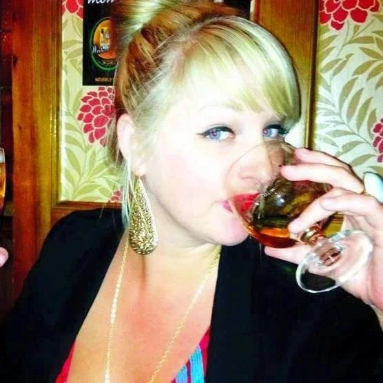A woman with blonde hair in a bun, wearing large earrings and a necklace, drinking from a glass of wine in a restaurant with floral wallpaper.