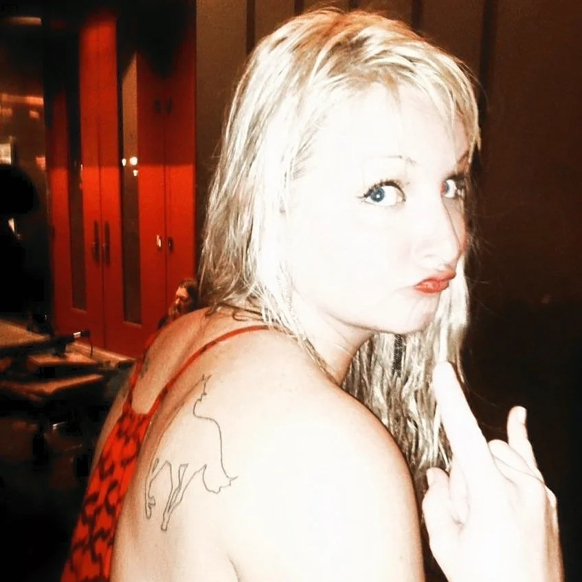 A woman with blonde hair and blue eyes, making a gesture with her middle finger, wearing a red and black patterned top, showing a tattoo of a lion on her shoulder, in an indoor setting with wooden cabinets in the background.