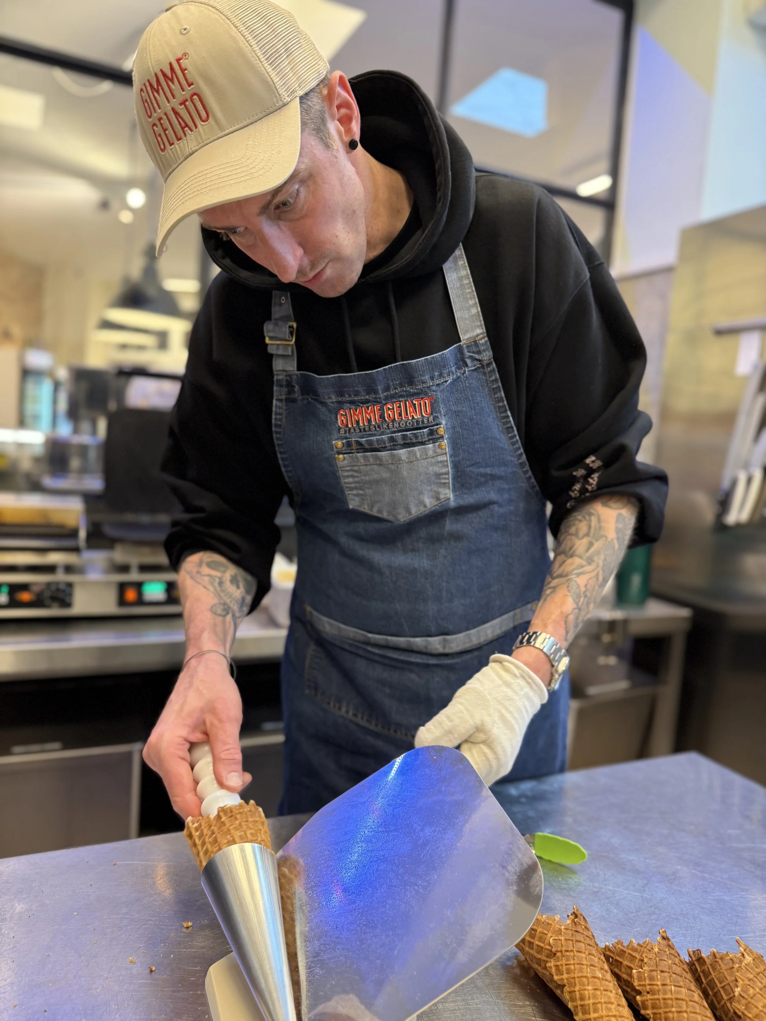Ein Mann in schwarzem Kapuzenpulli, blauer Latzhose und beigem Cap mit rotem Aufdruck 'GIMME GELATO' macht Eis in einer Eiswaffel in einer Eisdiele.
